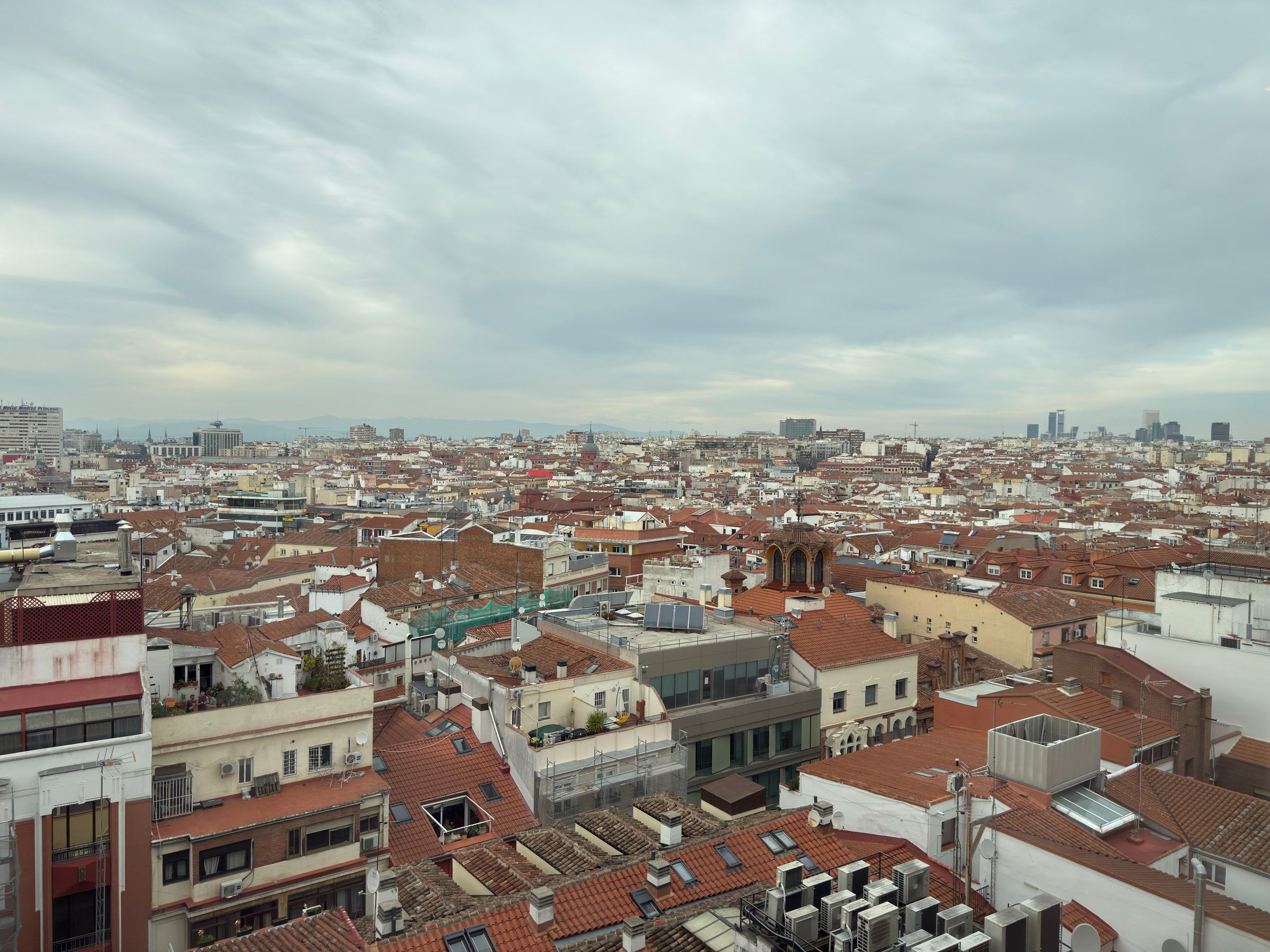 A wide view across a city of terracotta rooftops under a flat grey sky, aerials and roof terraces below and distant towers on the skyline