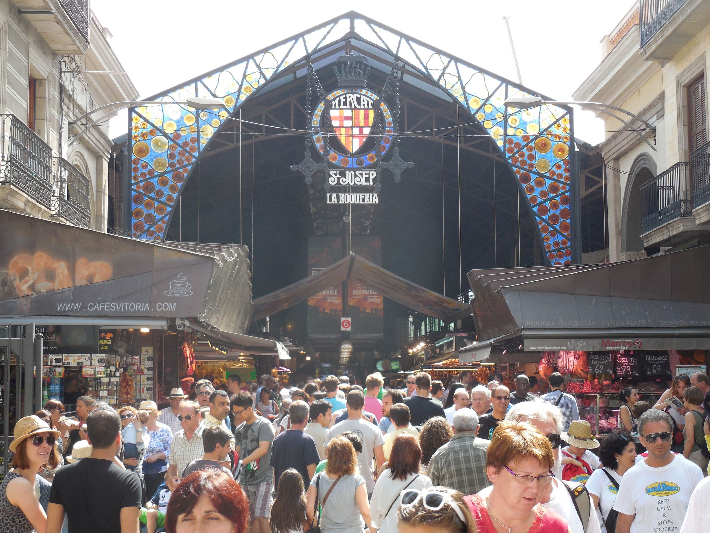 The iron entrance arch of the Sant Josep market under a stained-glass sign, a dense crowd passing beneath