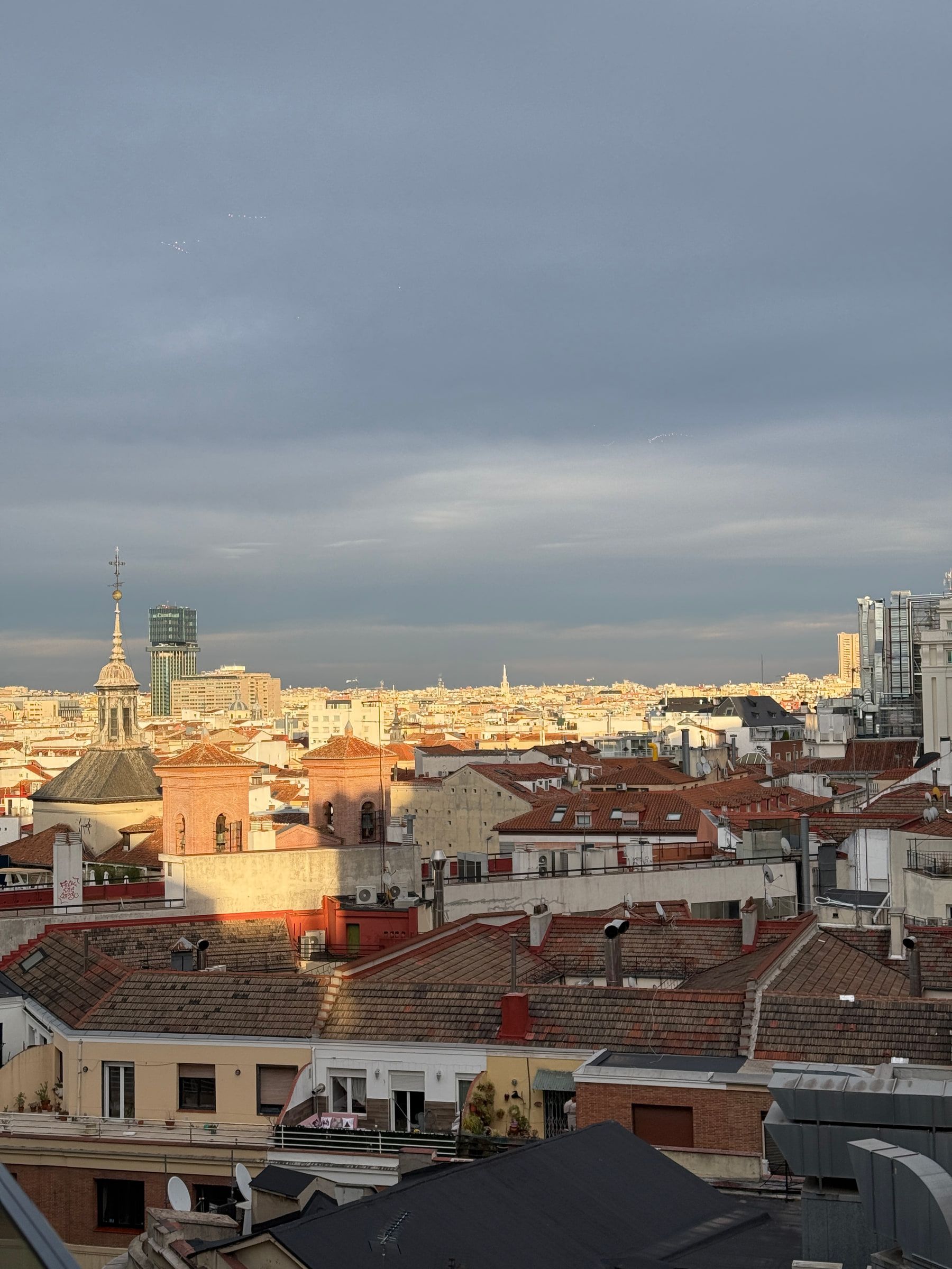 City rooftops catching low orange sun beneath a wide bank of cloud, a church dome and spire among them and towers on the far skyline