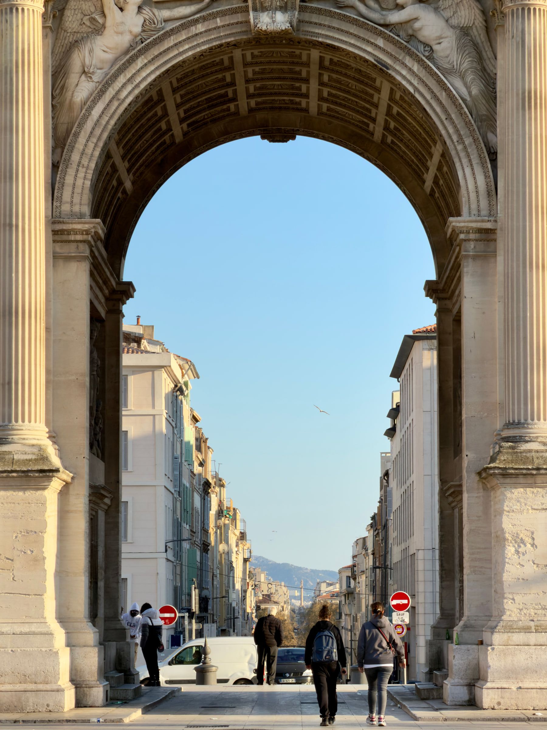 A tall stone triumphal arch framing a street that runs downhill between pale buildings toward distant hills, with people crossing beneath
