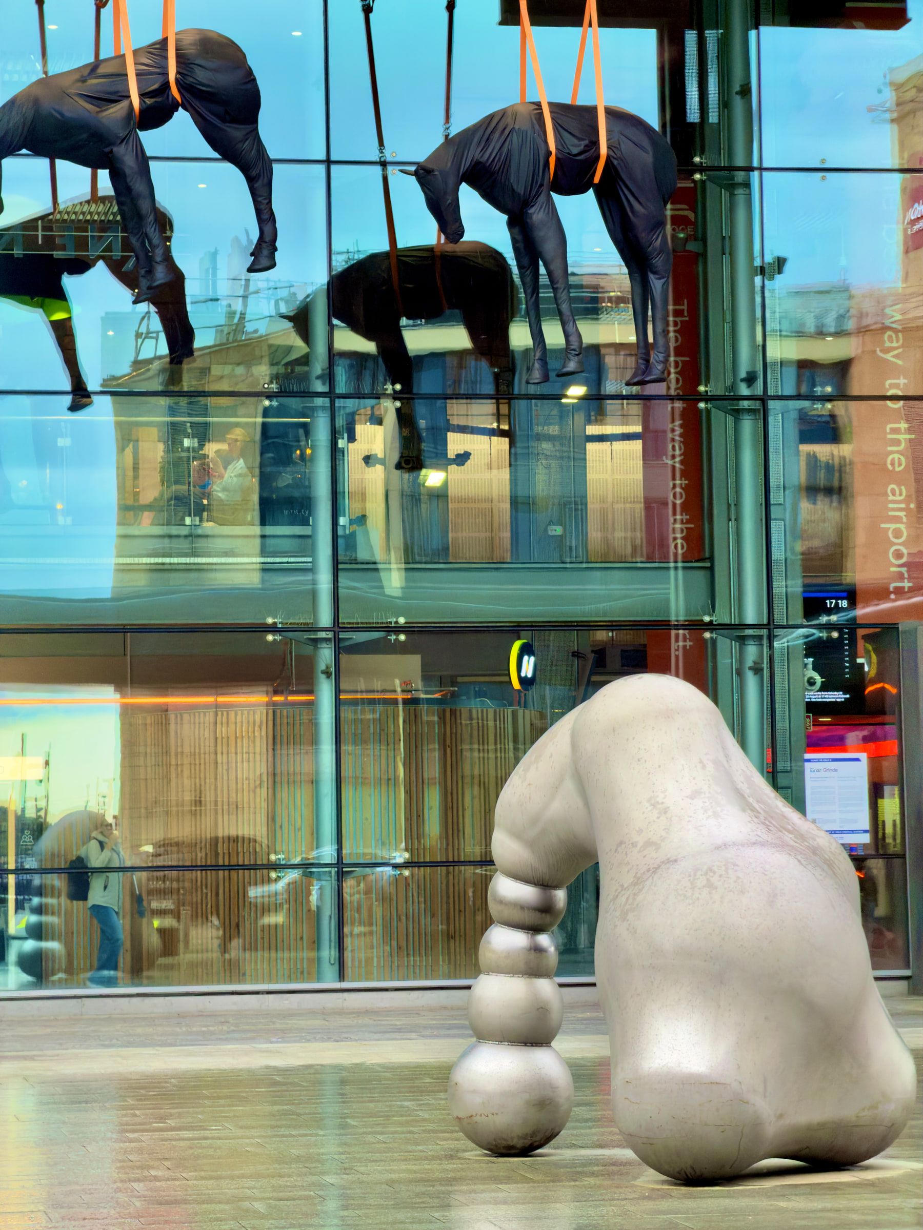 Dark figurative sculptures suspended above a glass entrance, with a large pale stone form standing on the pavement below
