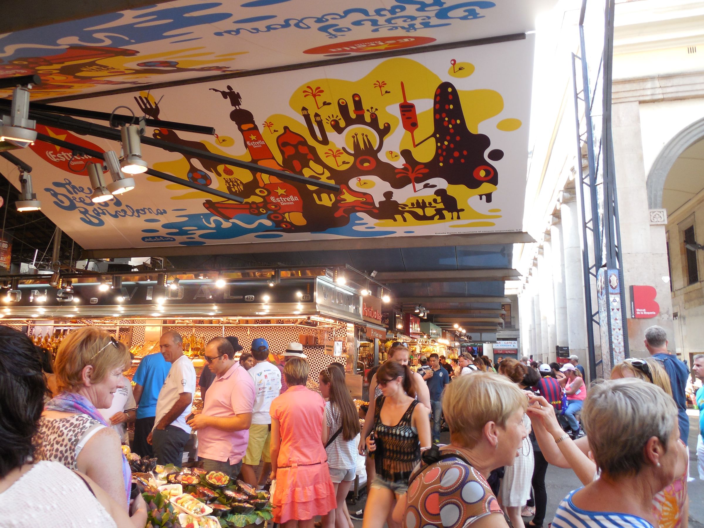Inside a covered market, a brightly painted mural ceiling above lit stalls and a crowd of shoppers