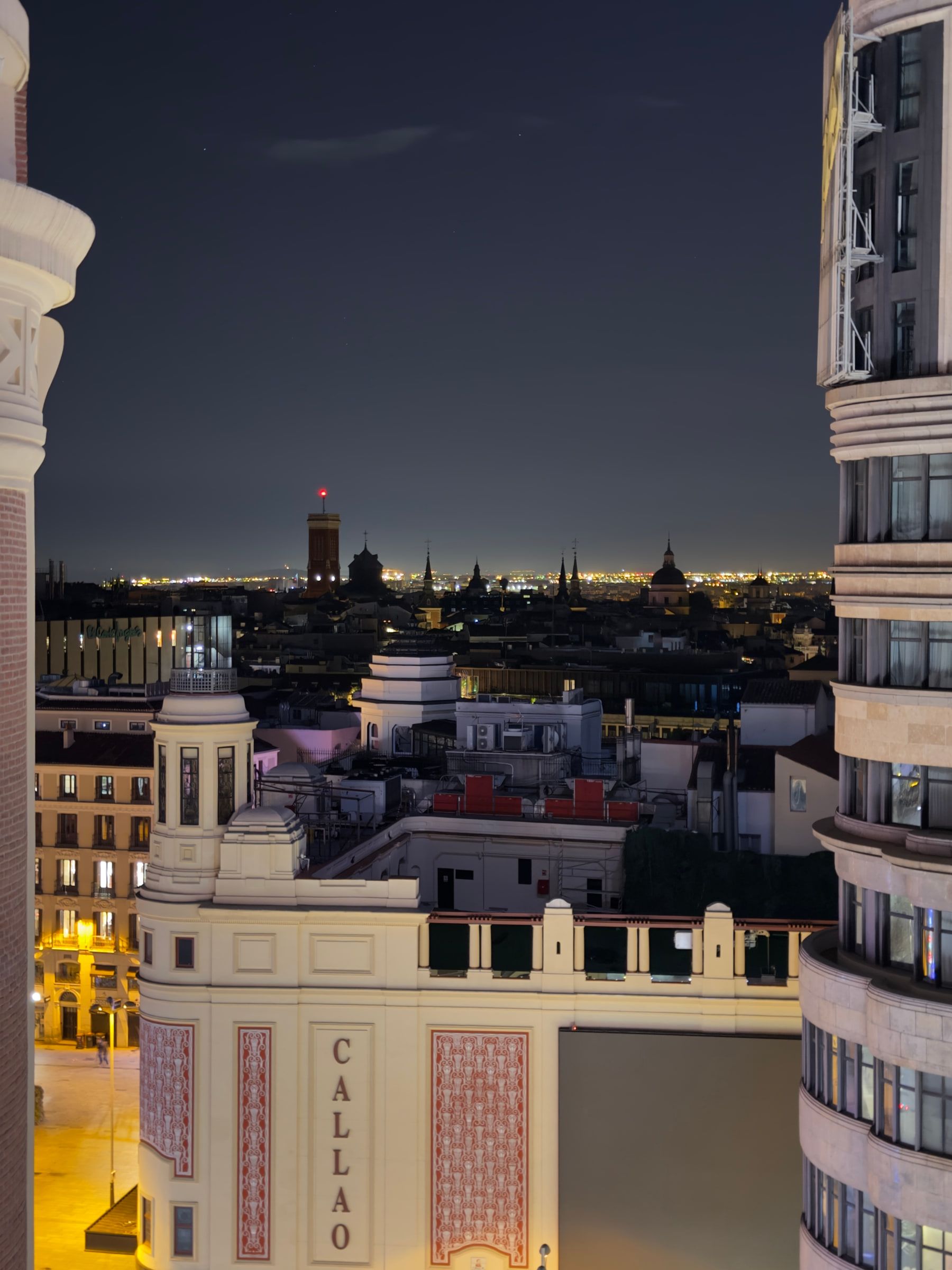 A night view between two tall buildings to a lit cinema front with CALLAO in vertical letters, the city's lights running to the horizon behind