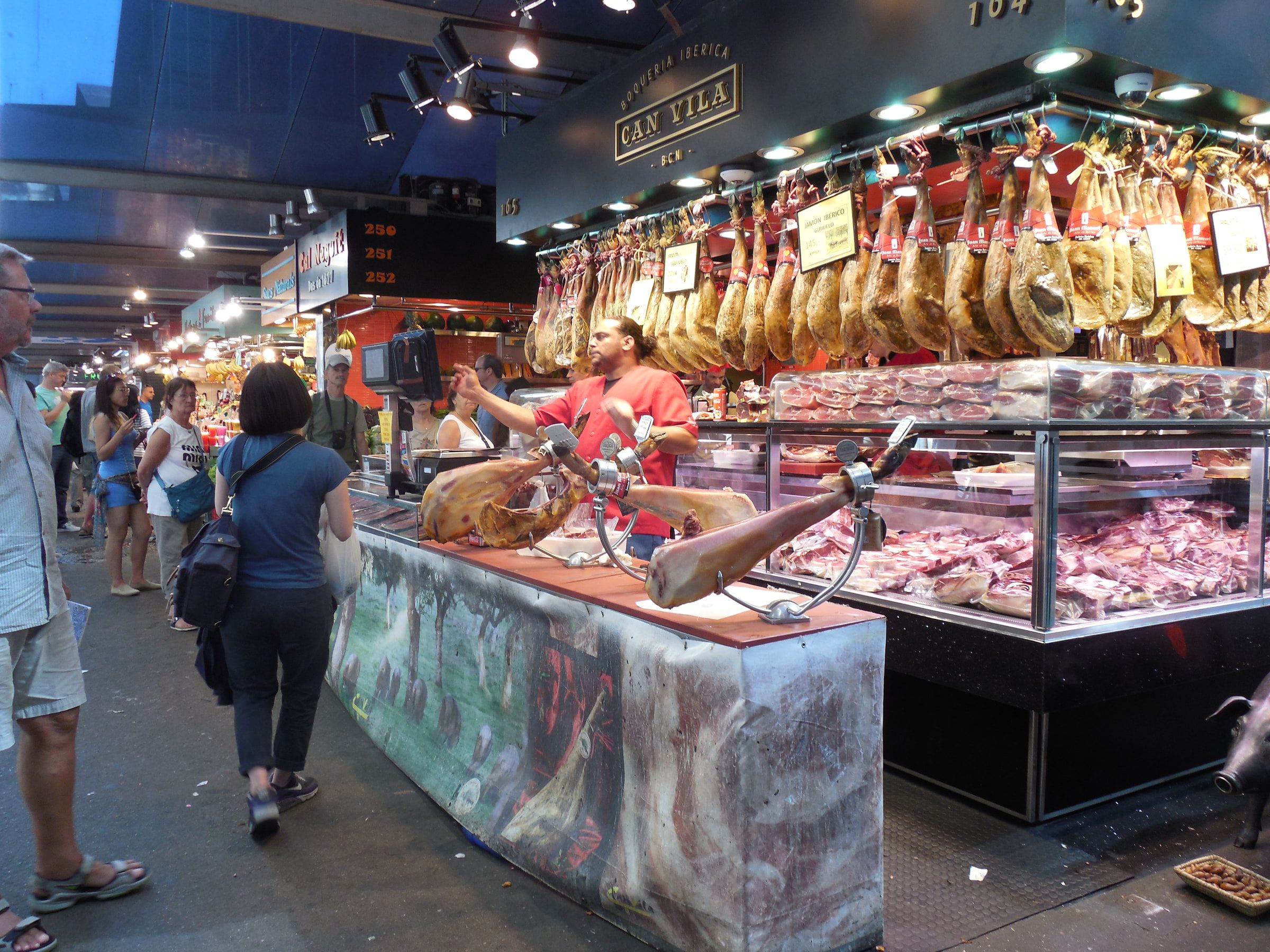A market counter hung with cured hams above a refrigerated case, a vendor working behind it