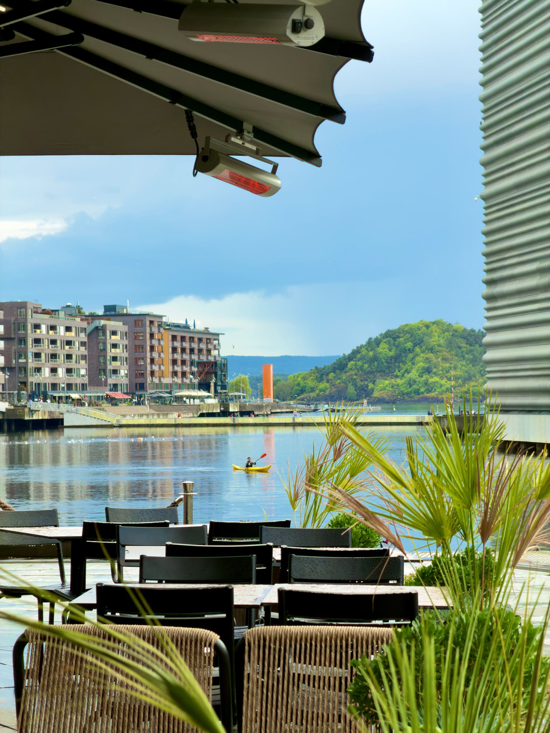 A waterside restaurant terrace with patio heaters and folded umbrellas, looking across the harbour to apartment blocks and a kayaker