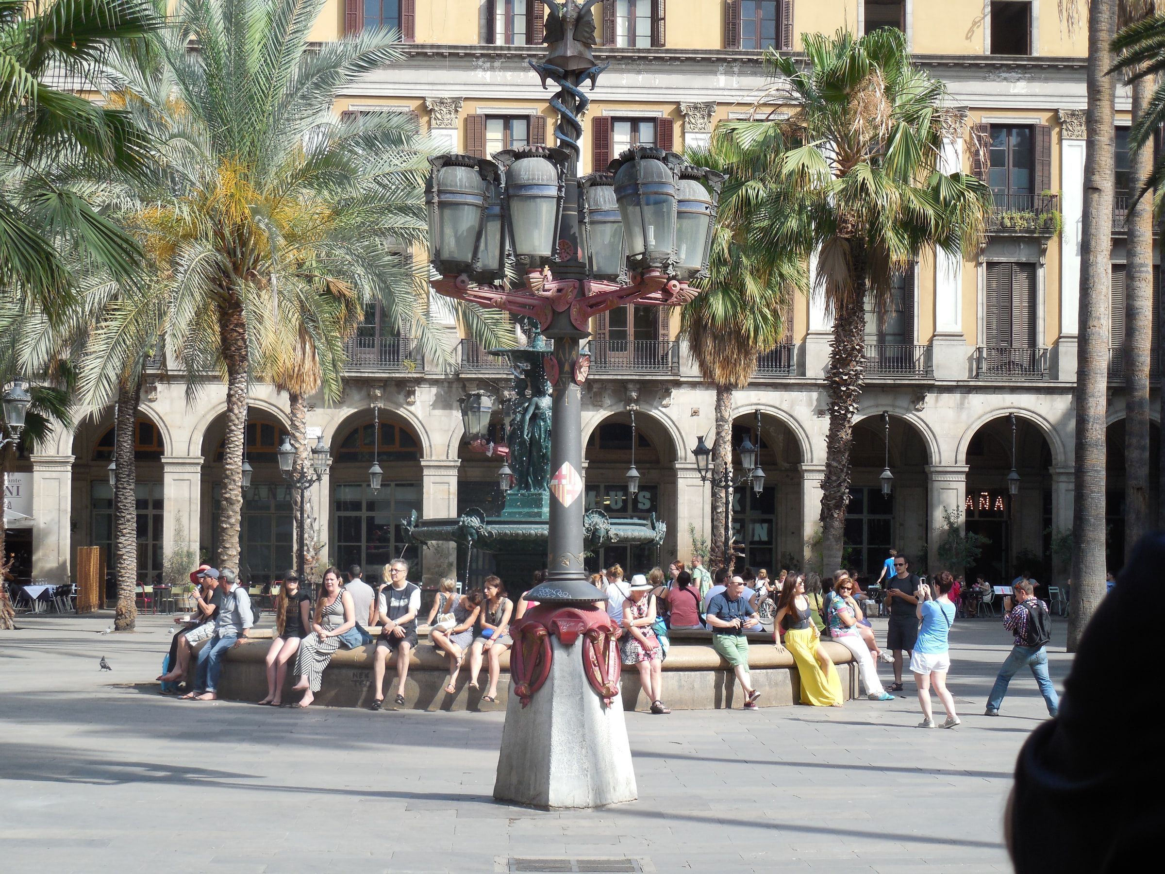 An arcaded square with tall palms and an ornate iron lamppost, people sitting along a low bench