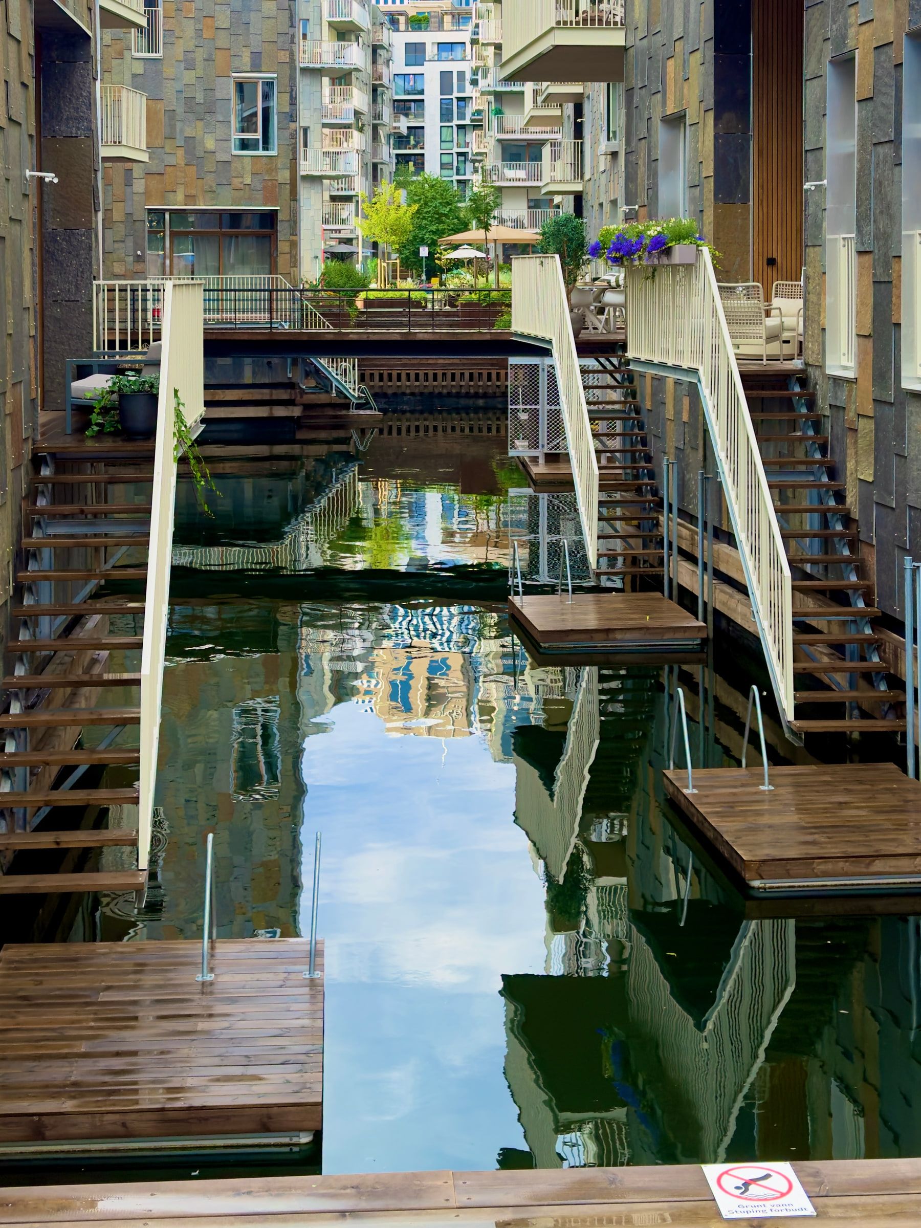 A narrow canal between apartment blocks, crossed by timber footbridges and decks that reflect in the still water