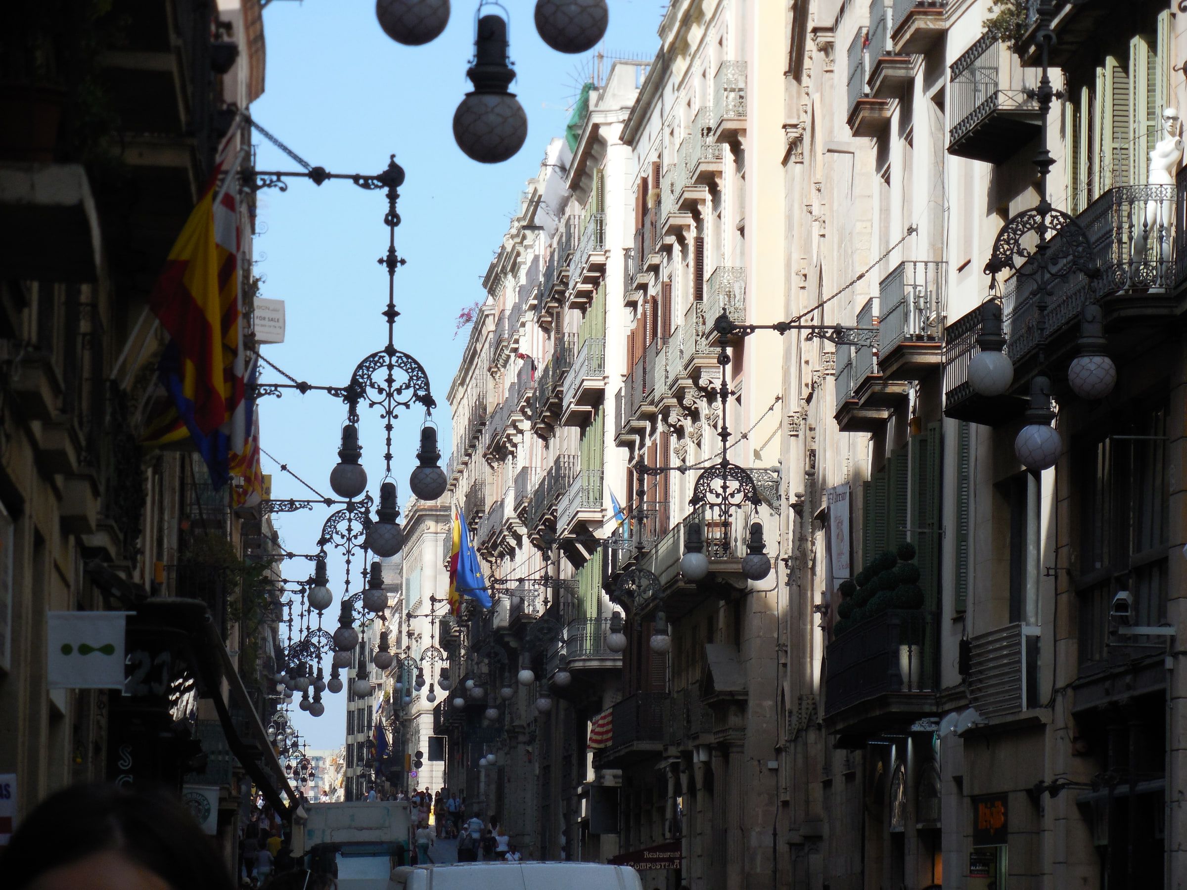 A narrow street strung with round hanging lamps between balconied buildings, a flag overhead