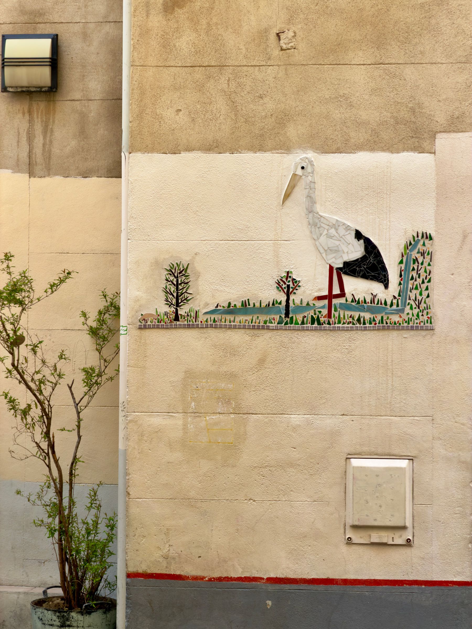 A mosaic panel of a white stork standing in grass, set into a plain rendered wall beside a young tree
