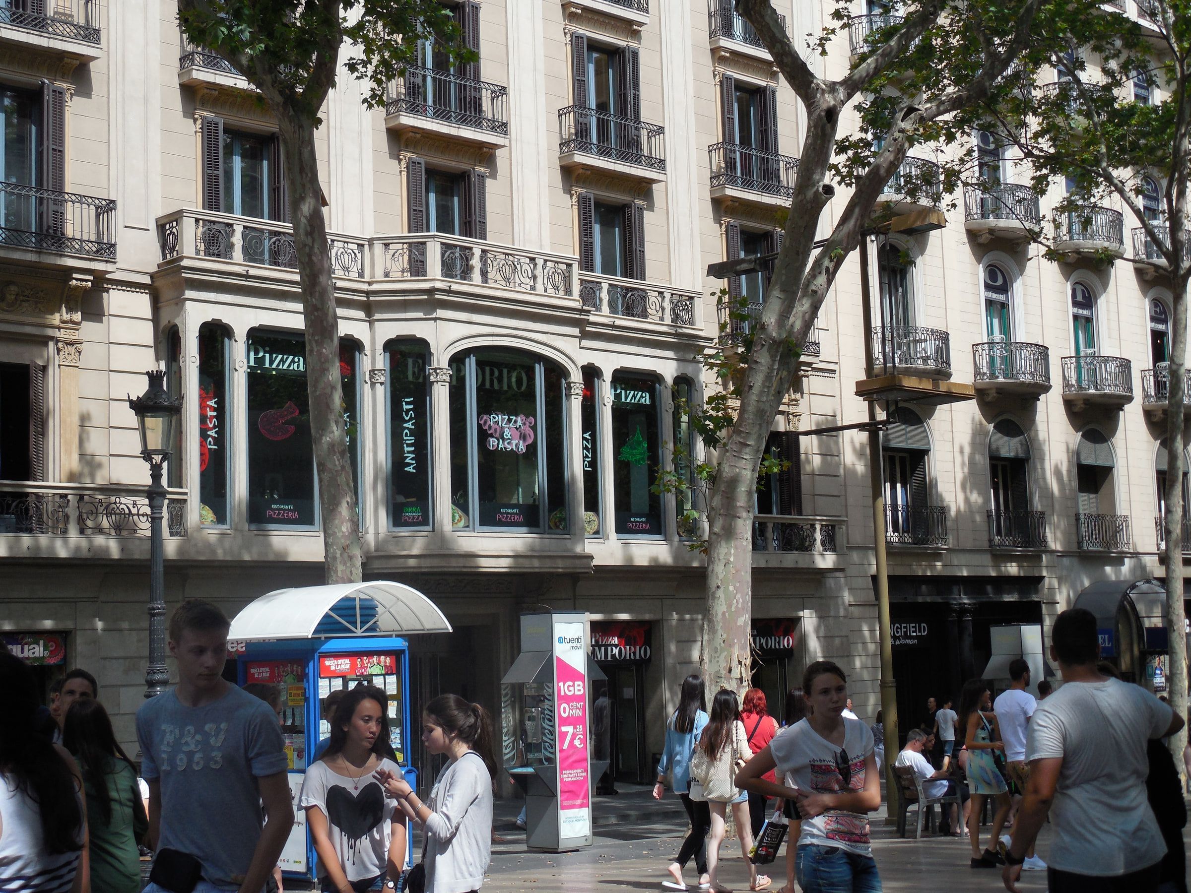 A broad street lined with plane trees and shopfronts in hard sun, people walking the pavement