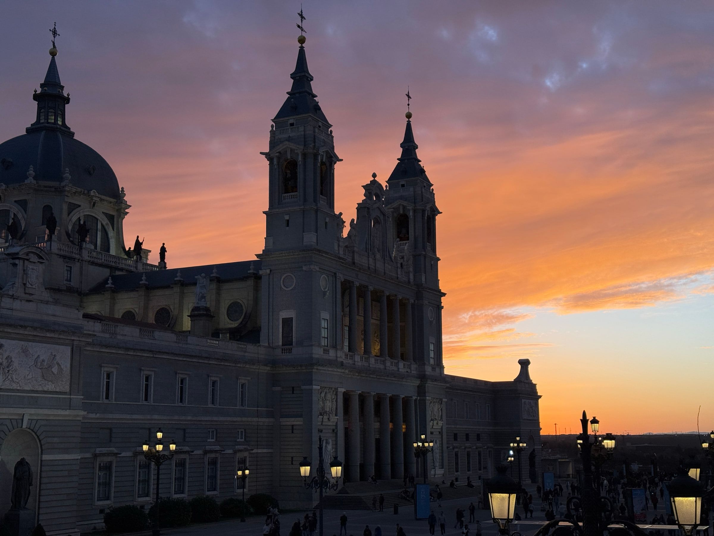A domed cathedral standing in silhouette against a sky burning orange and pink, lamps lit in the plaza below and people crossing it