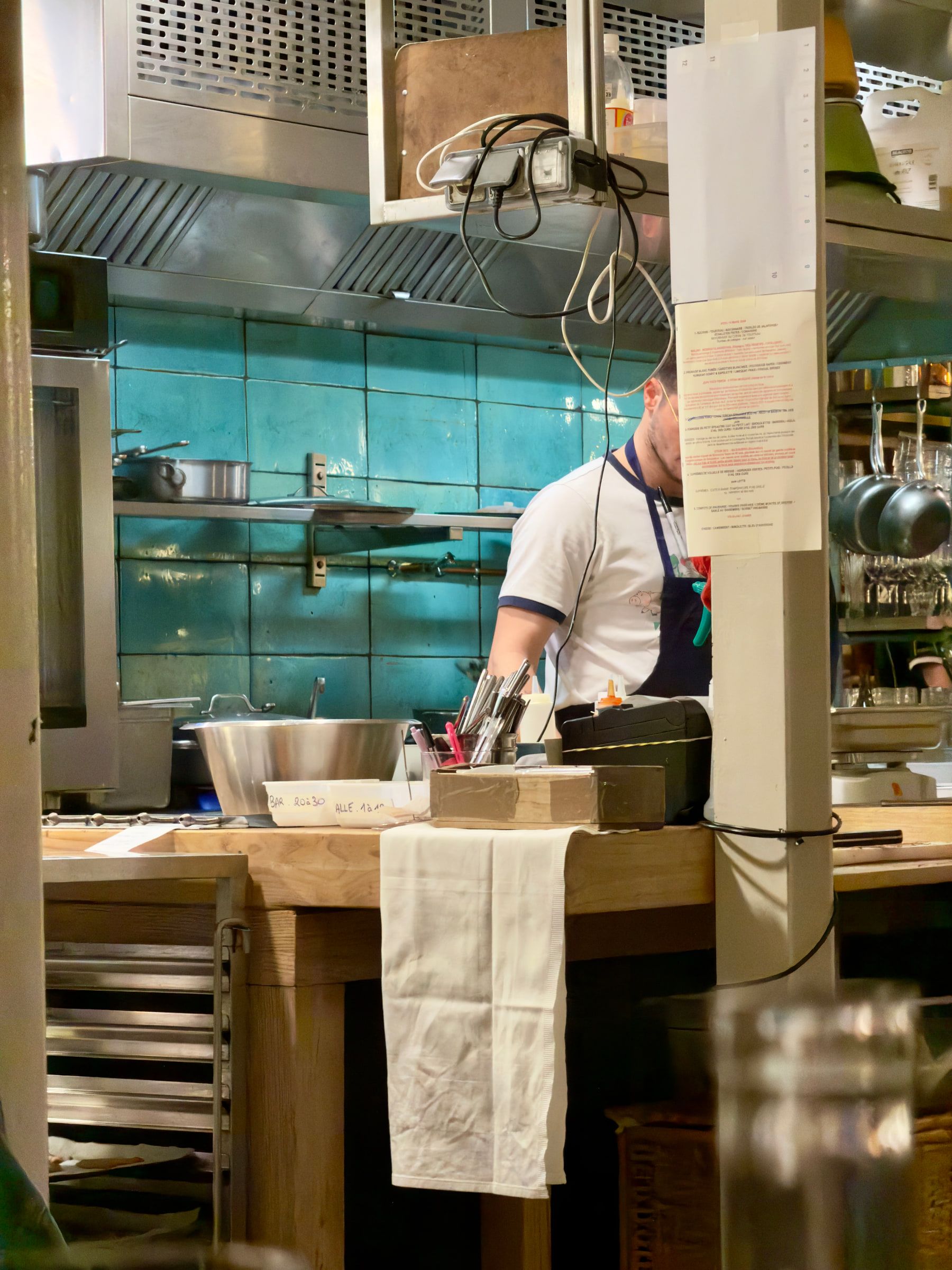 A cook working behind a small restaurant counter, turquoise tiles and hanging pans behind him, a folded cloth over the counter edge