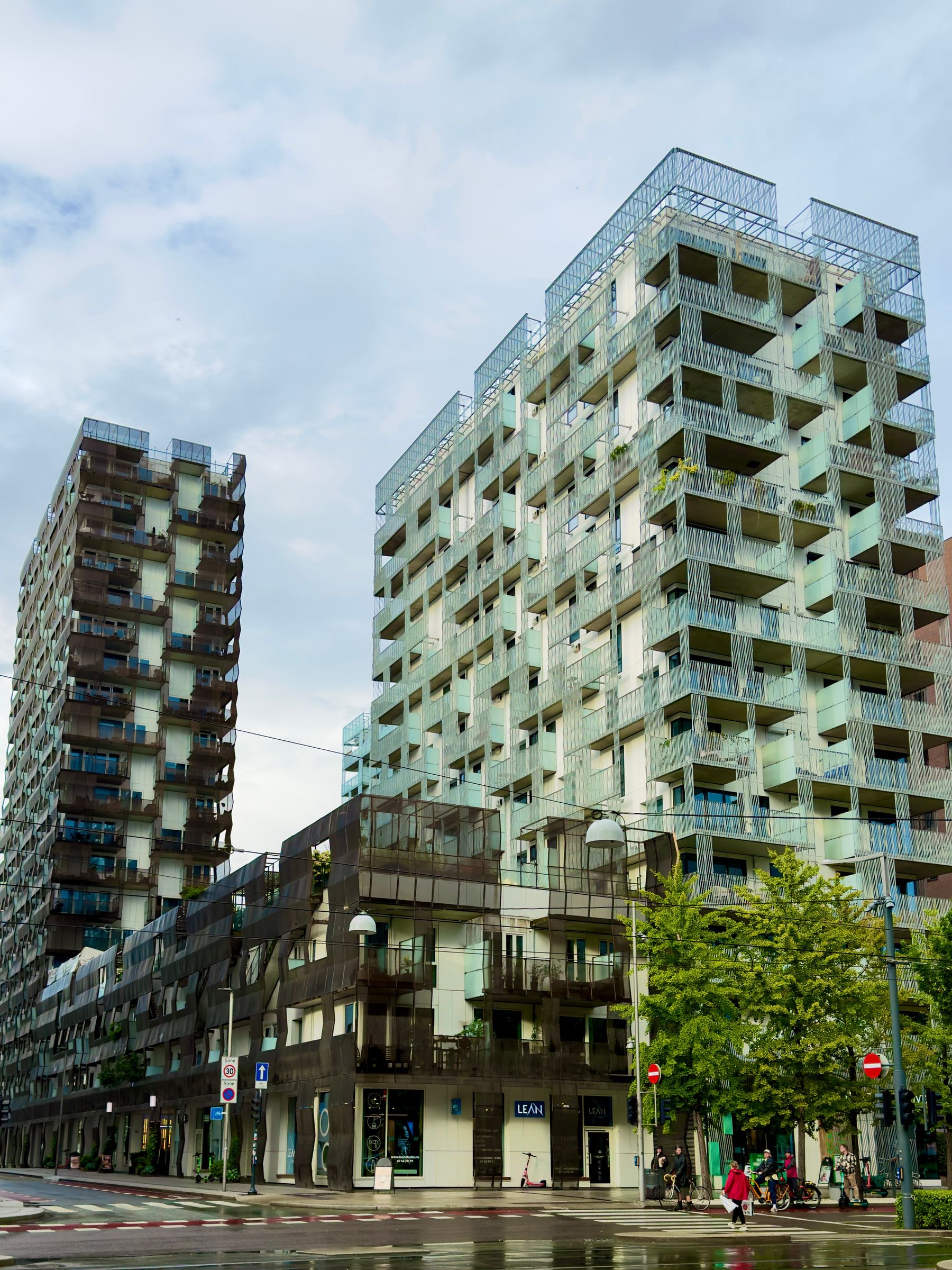 Two tall residential towers with stacked balconies rising over a wet street with trees and shopfronts