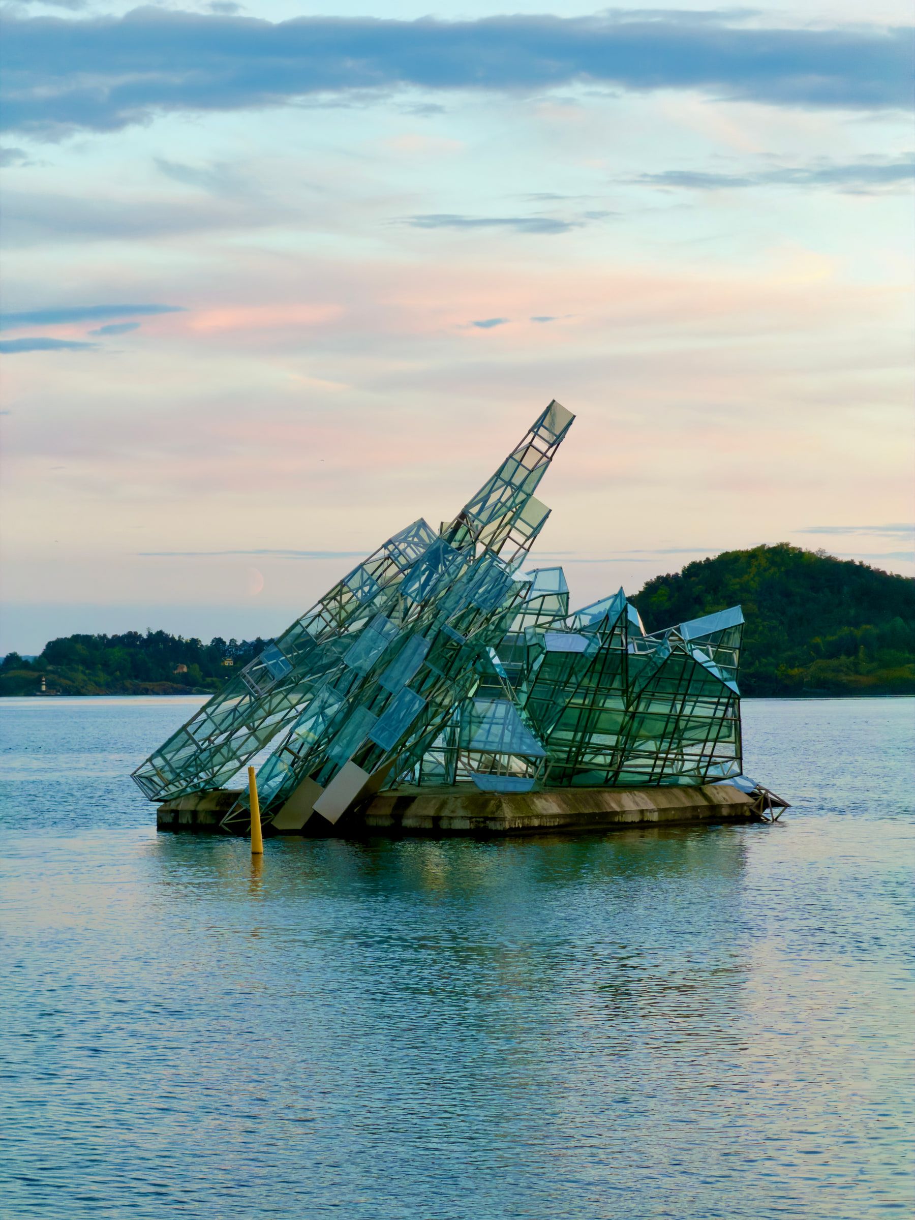 An angular glass-and-steel structure tilted in shallow coastal water at dusk, a low island behind it and a pink sky above