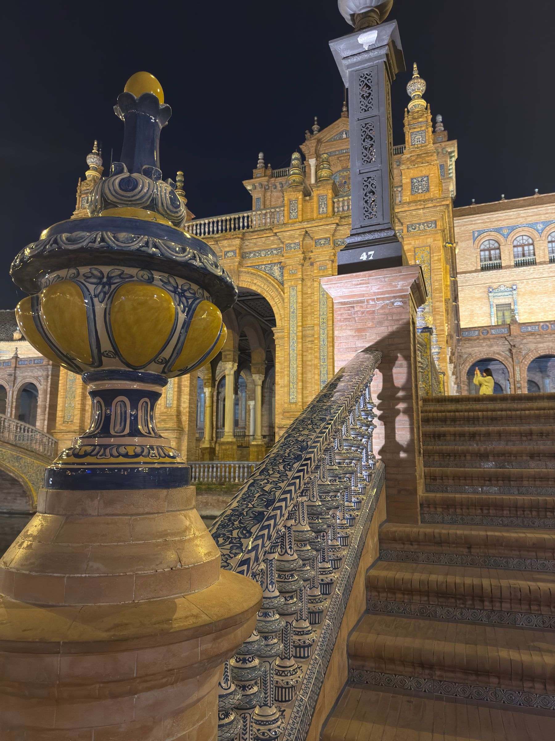 A painted blue and yellow ceramic urn on a tiled balustrade at night, beside a flight of steps and a lamp post, a tiled facade rising behind