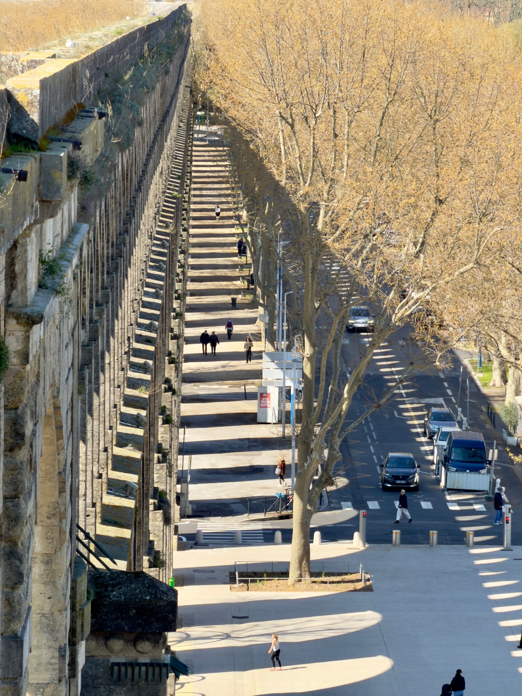 A long stone aqueduct running beside a tree-lined boulevard in low sun, its arches throwing repeated shadows across the pavement