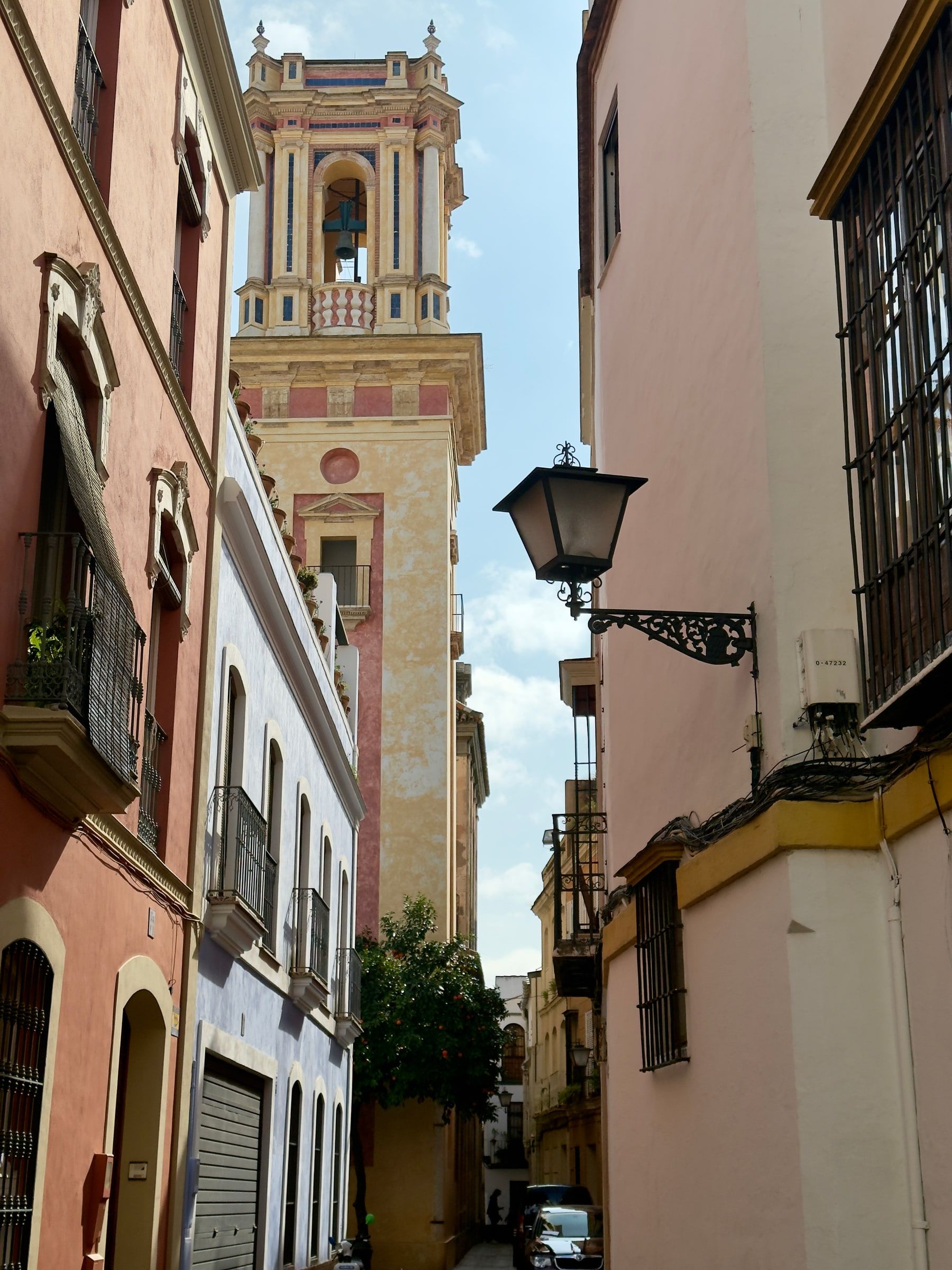 A narrow street of pink and ochre houses with an iron lantern bracketed to a wall, an ornate baroque bell tower closing the far end