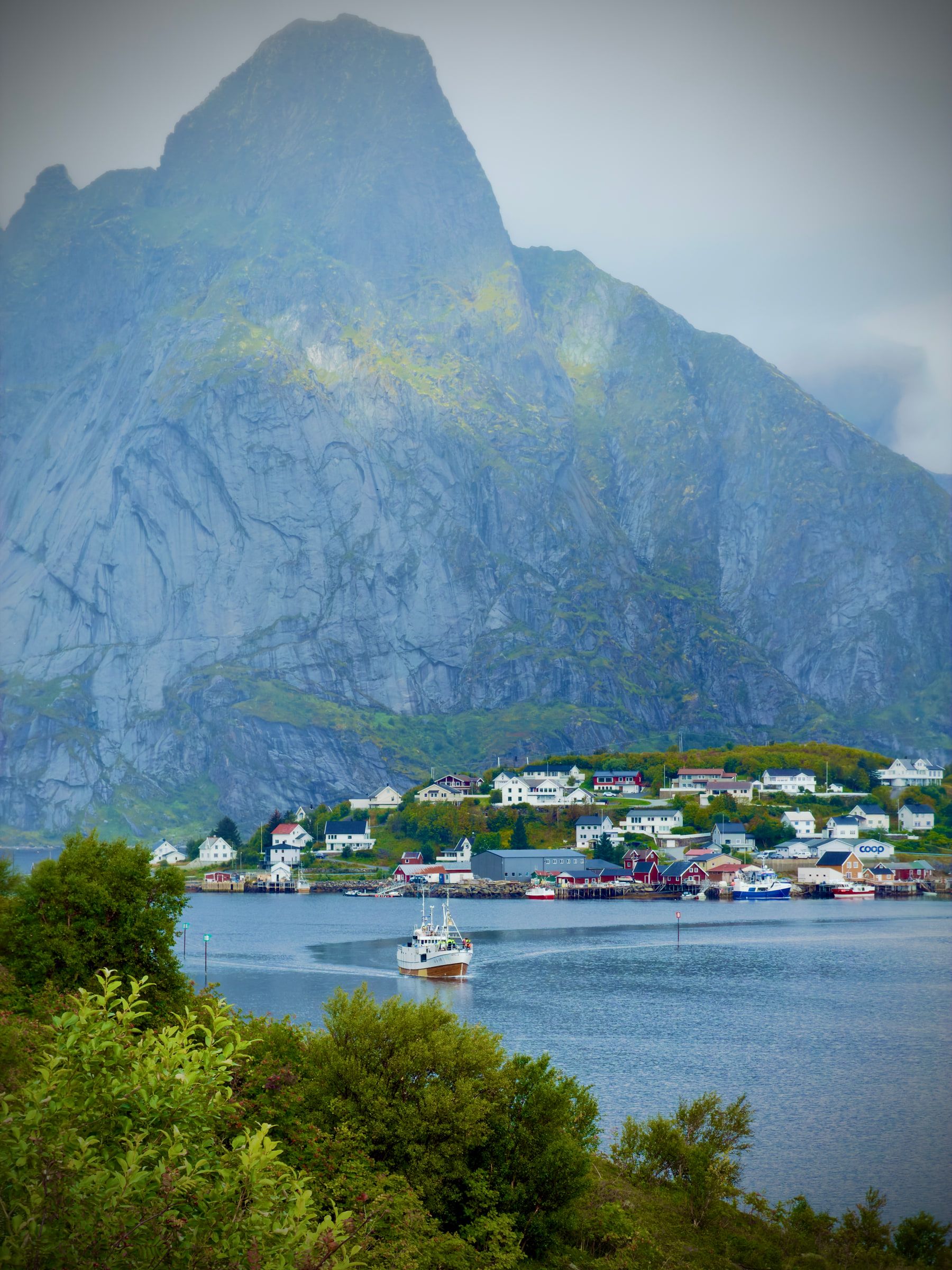 A sharp granite peak streaked with green rising above a shoreline village of white houses and moored boats