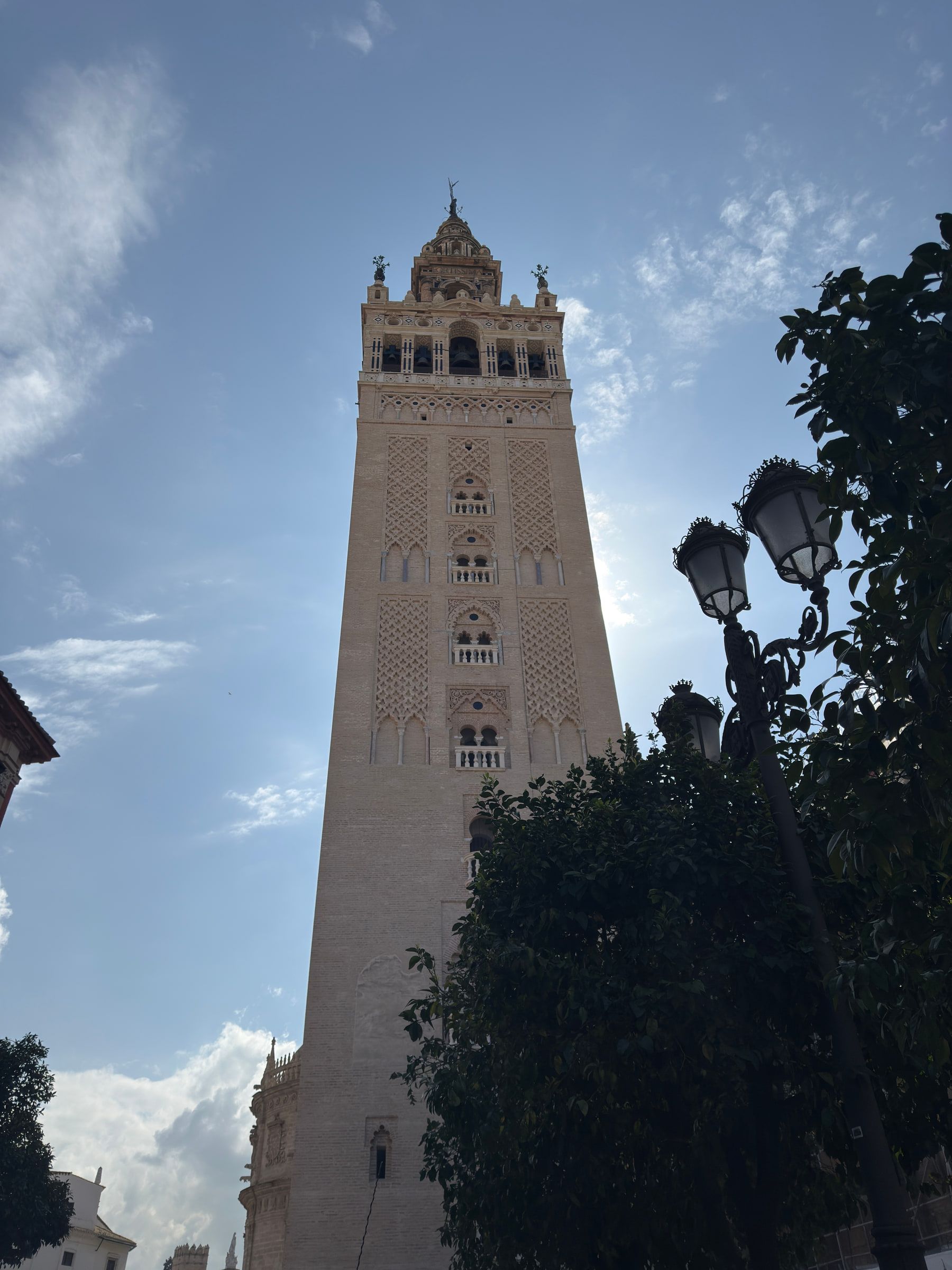 A tall square bell tower of pale stone and brick tracery photographed steeply from below against a blue sky, an ornate street lamp and dark foliage at the edge of the frame
