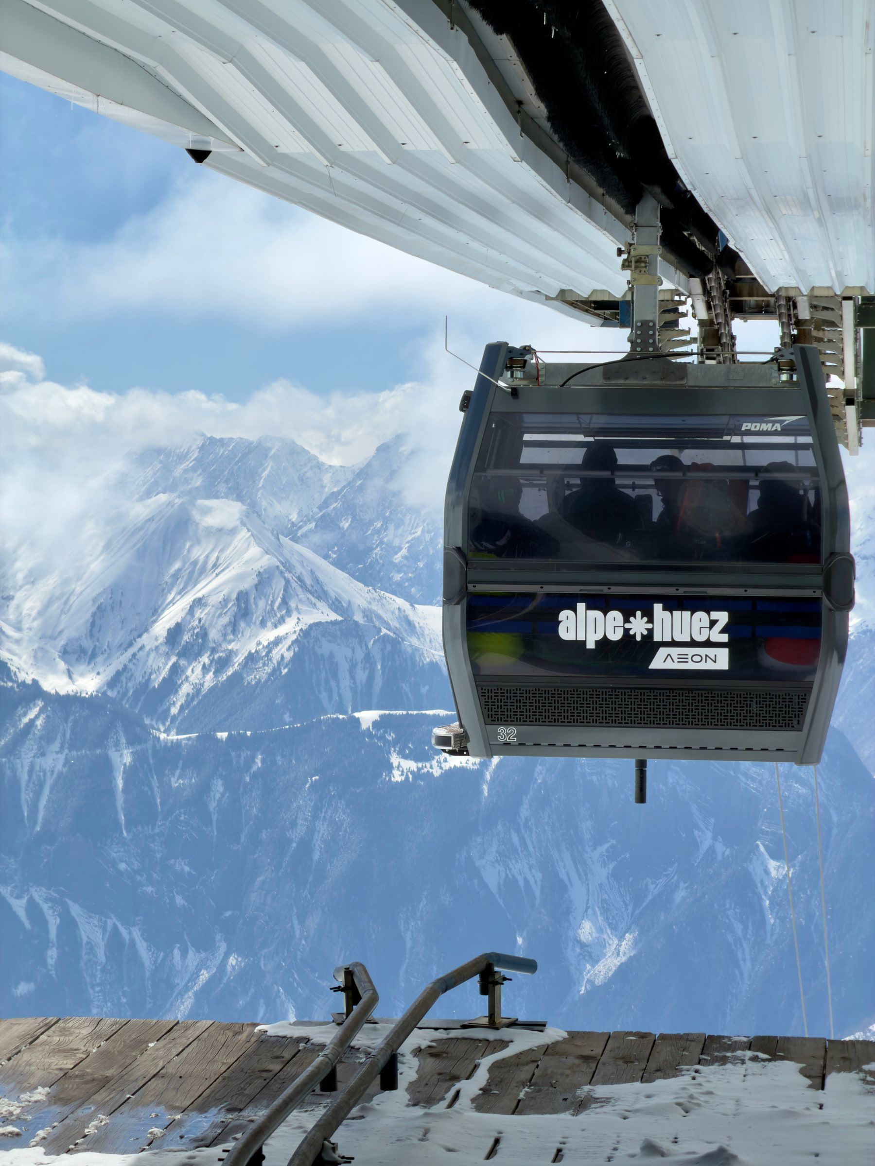 A gondola cabin marked Alpe d'Huez passing directly overhead, snowy peaks visible beyond it
