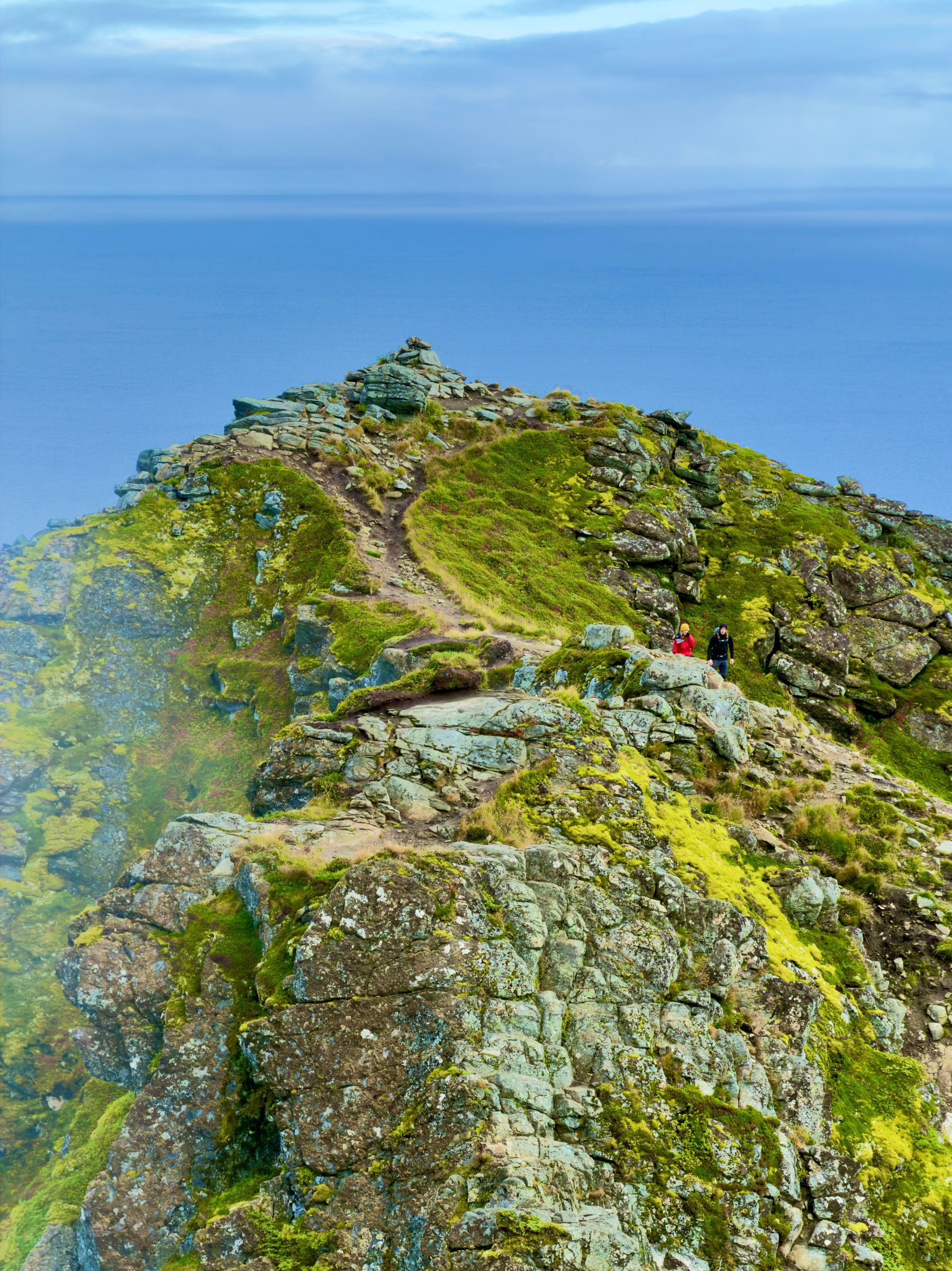 A rocky summit ridge dropping away to blue sea far below, with a single figure in a red jacket sitting near the top