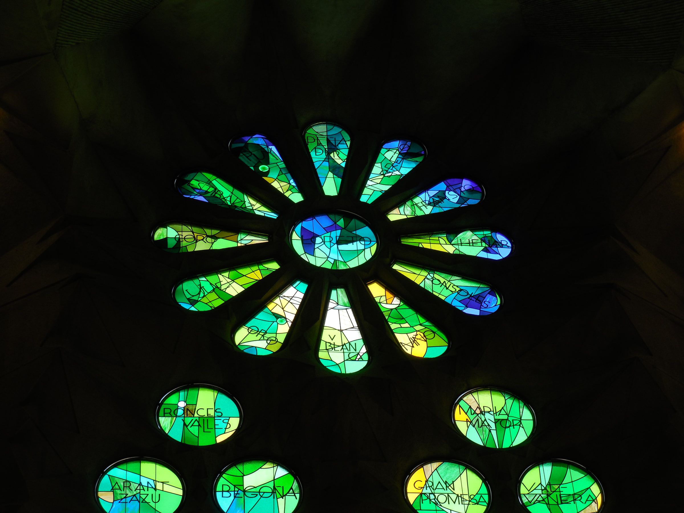 A circular stained-glass oculus in greens and blues set into a dark vault, smaller roundels beneath it