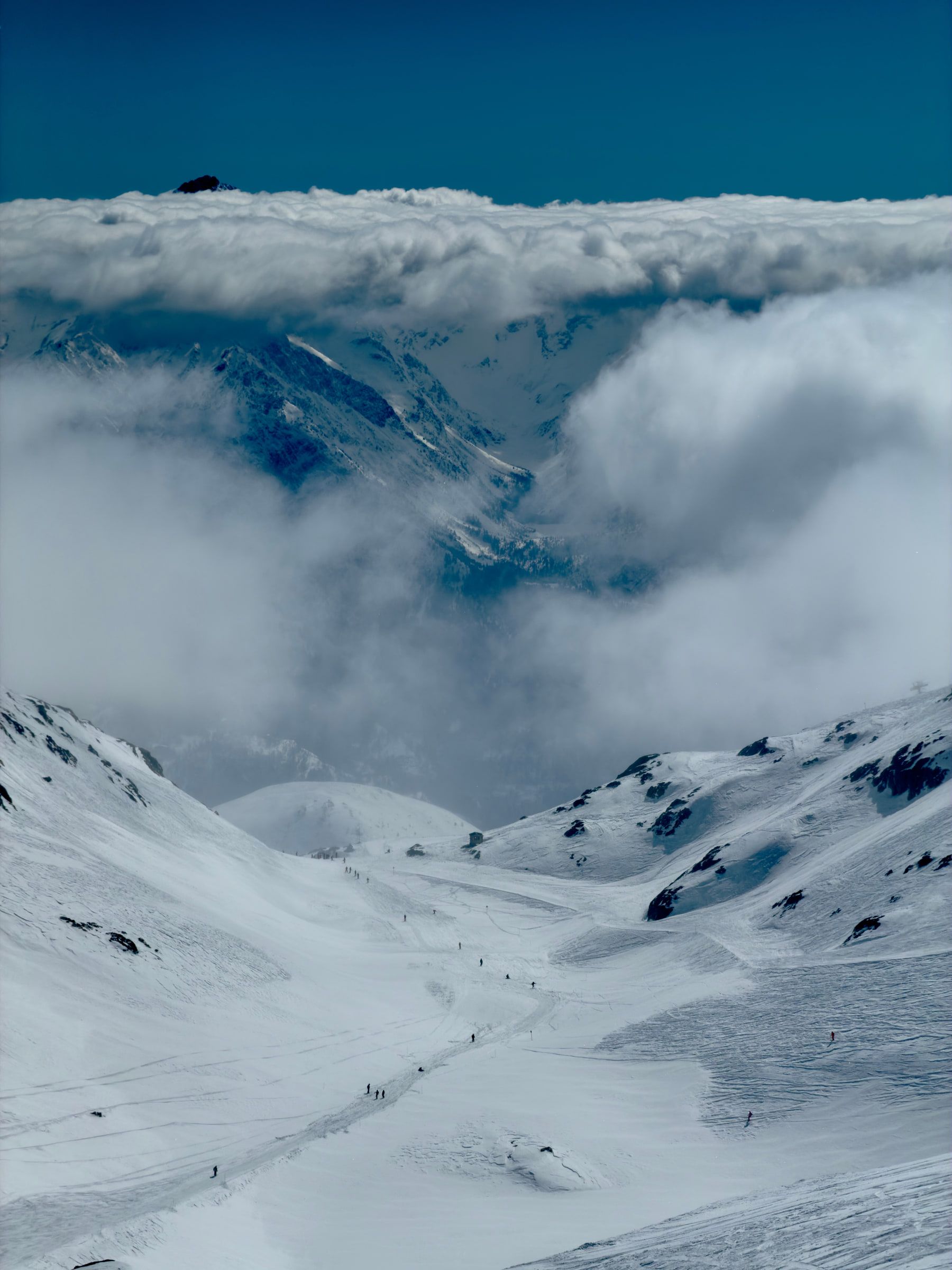 A piste running down toward a valley completely filled with cloud, dark rock breaking through above it