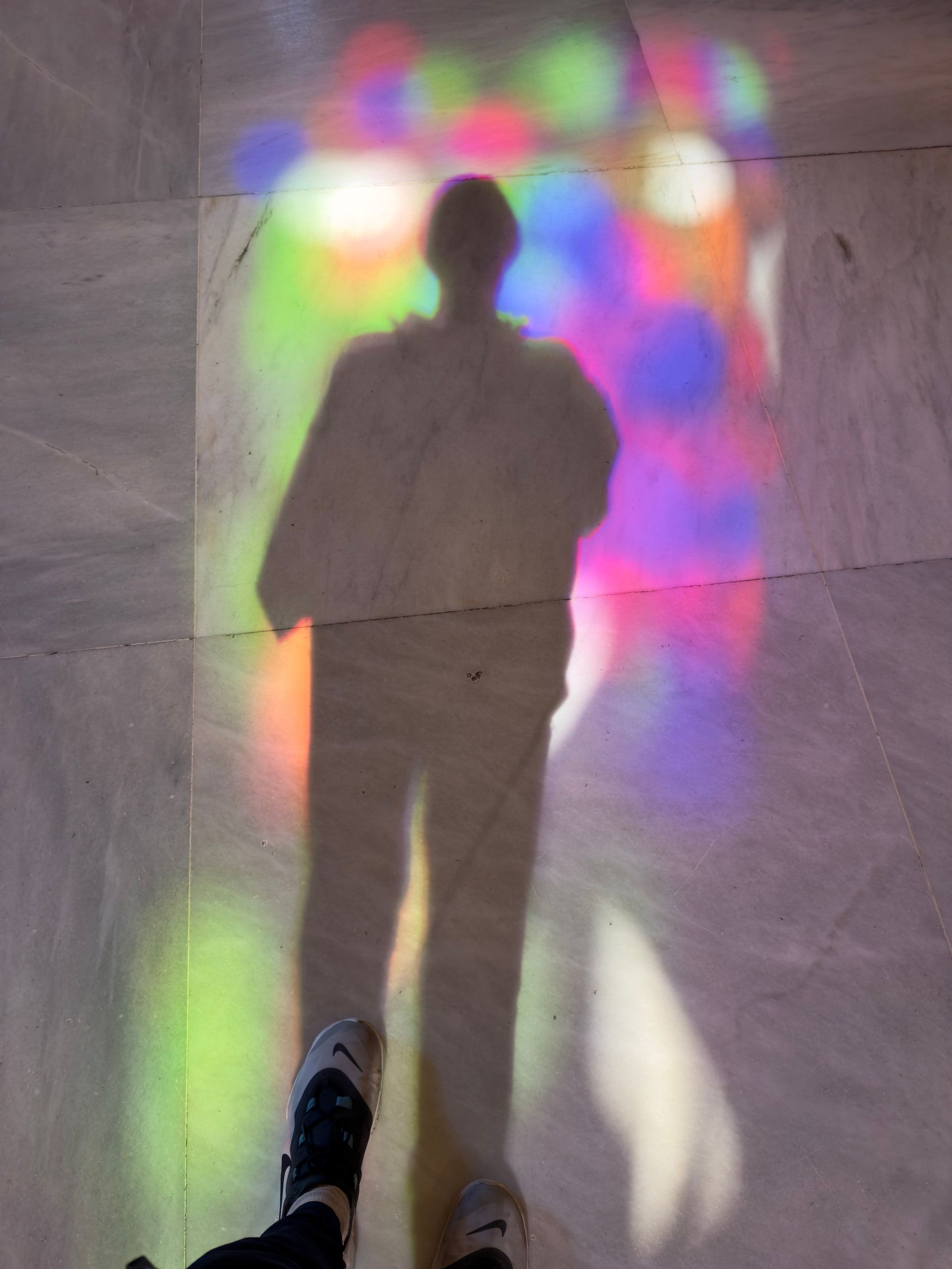 The photographer's long shadow cast on a stone floor washed with green, pink and blue light from stained glass, their shoes at the bottom of the frame