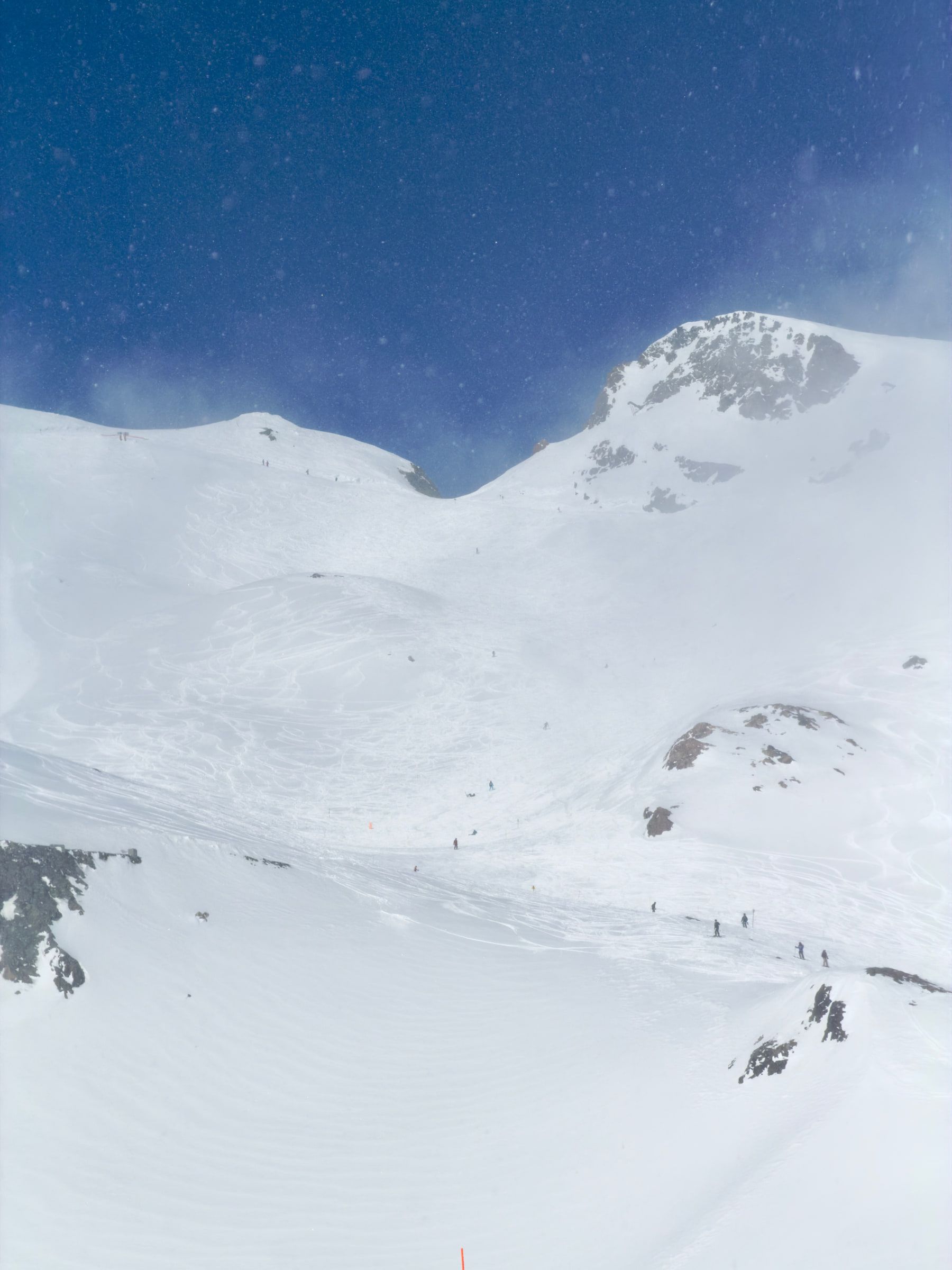 Wind-blown snow streaming across a broad white summit dome under a dark blue sky, skiers reduced to specks