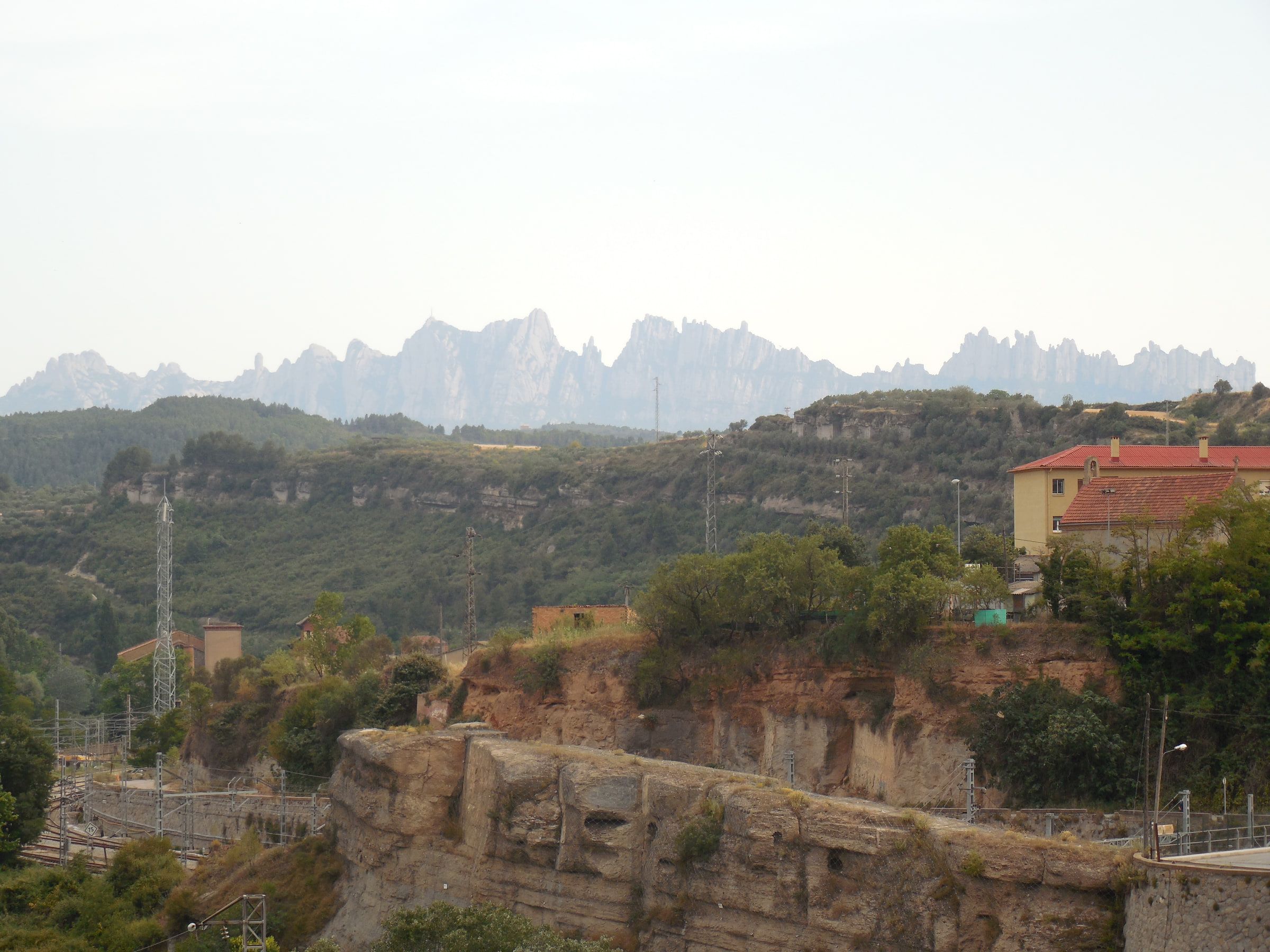 A distant serrated rock massif rising over wooded hills, seen past scrub and rooftops