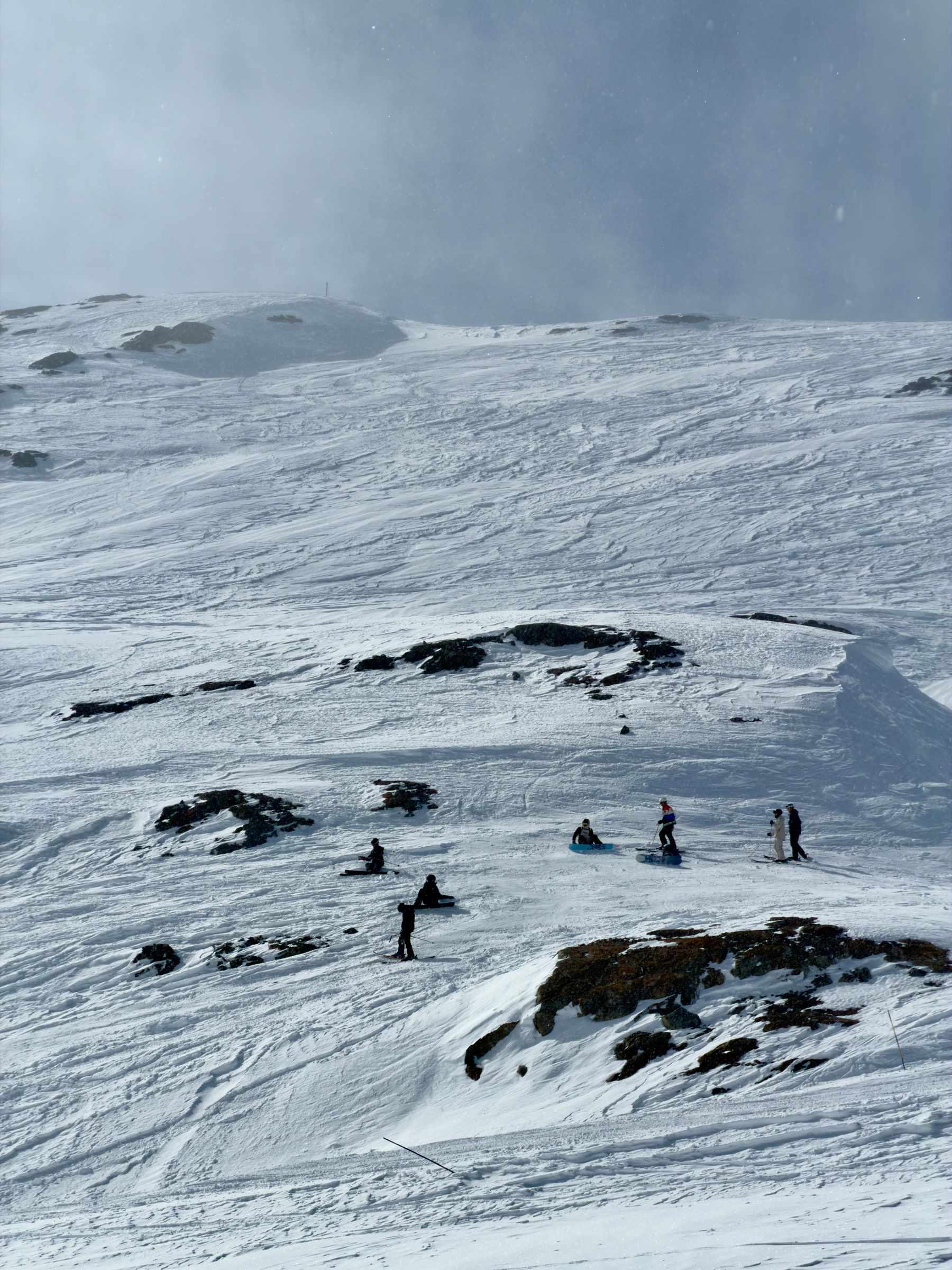 Skiers spread across a wide open snow slope with dark rock outcrops, in flat grey light