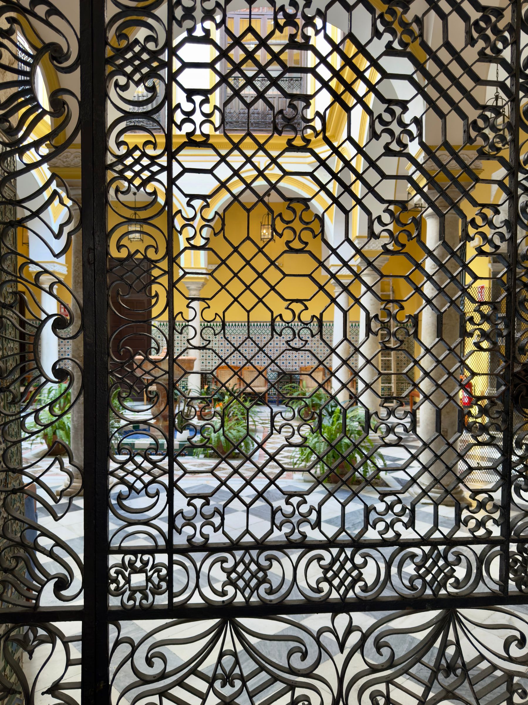 A bright yellow courtyard with arches, tiled dado and potted plants, seen through the dense scrollwork of a tall wrought-iron gate