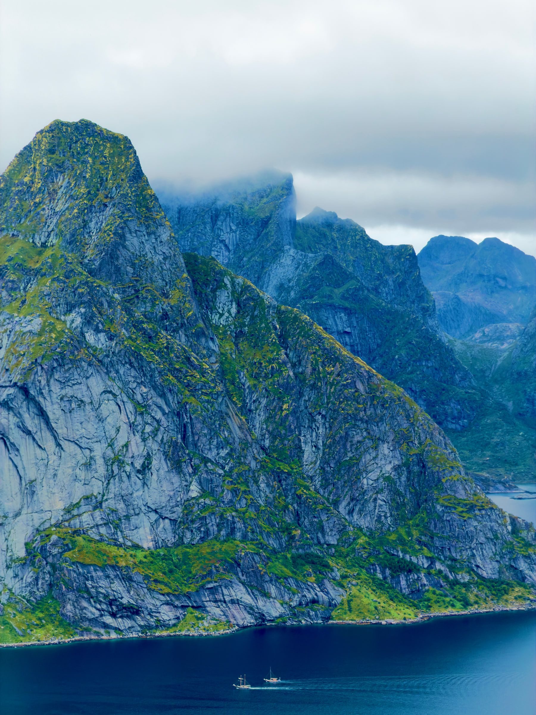 A steep mountain wall dropping straight into a deep blue fjord, its summit covered by cloud
