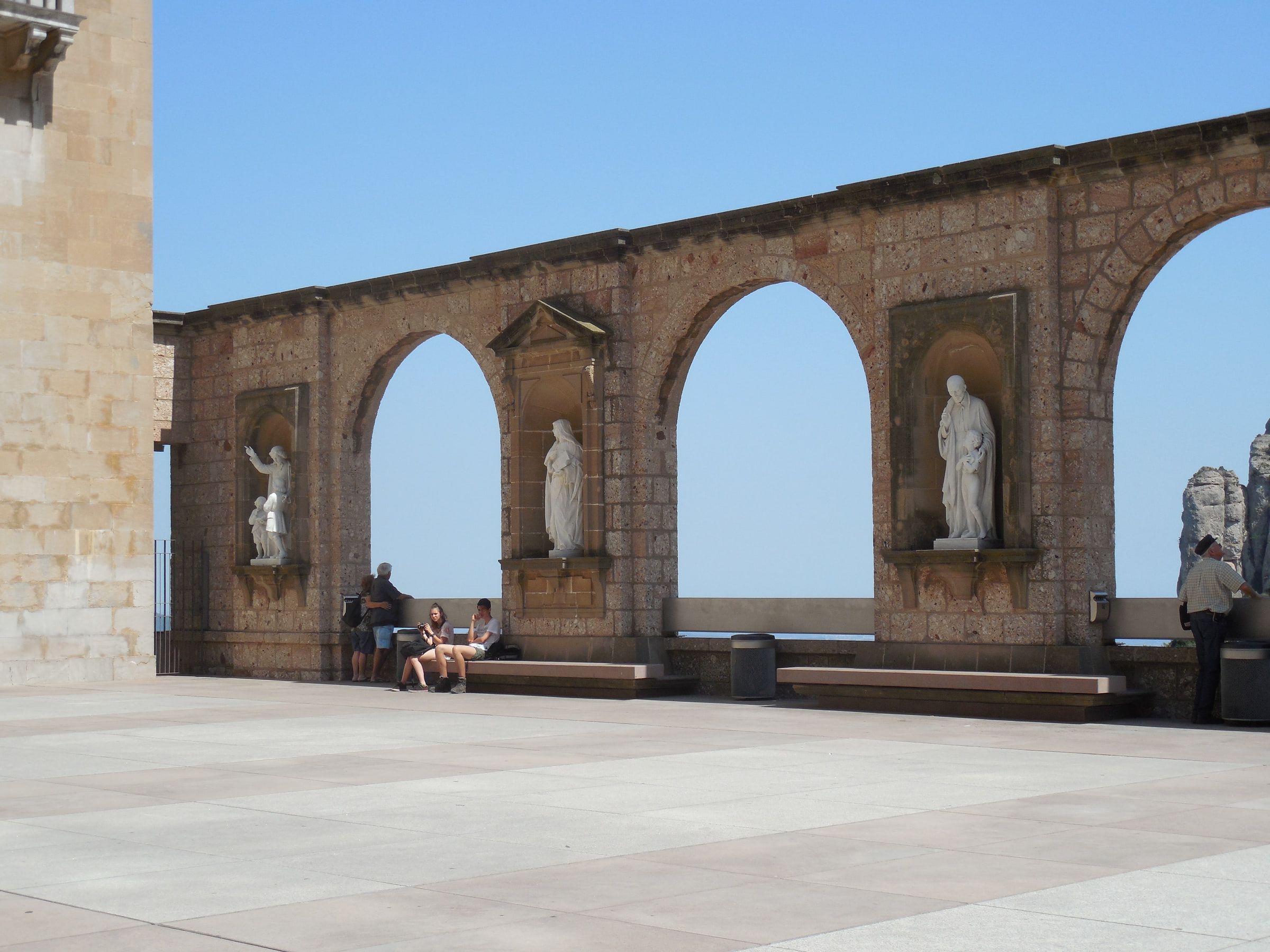 An arcaded stone terrace with statues standing in its niches, two people sitting on a bench