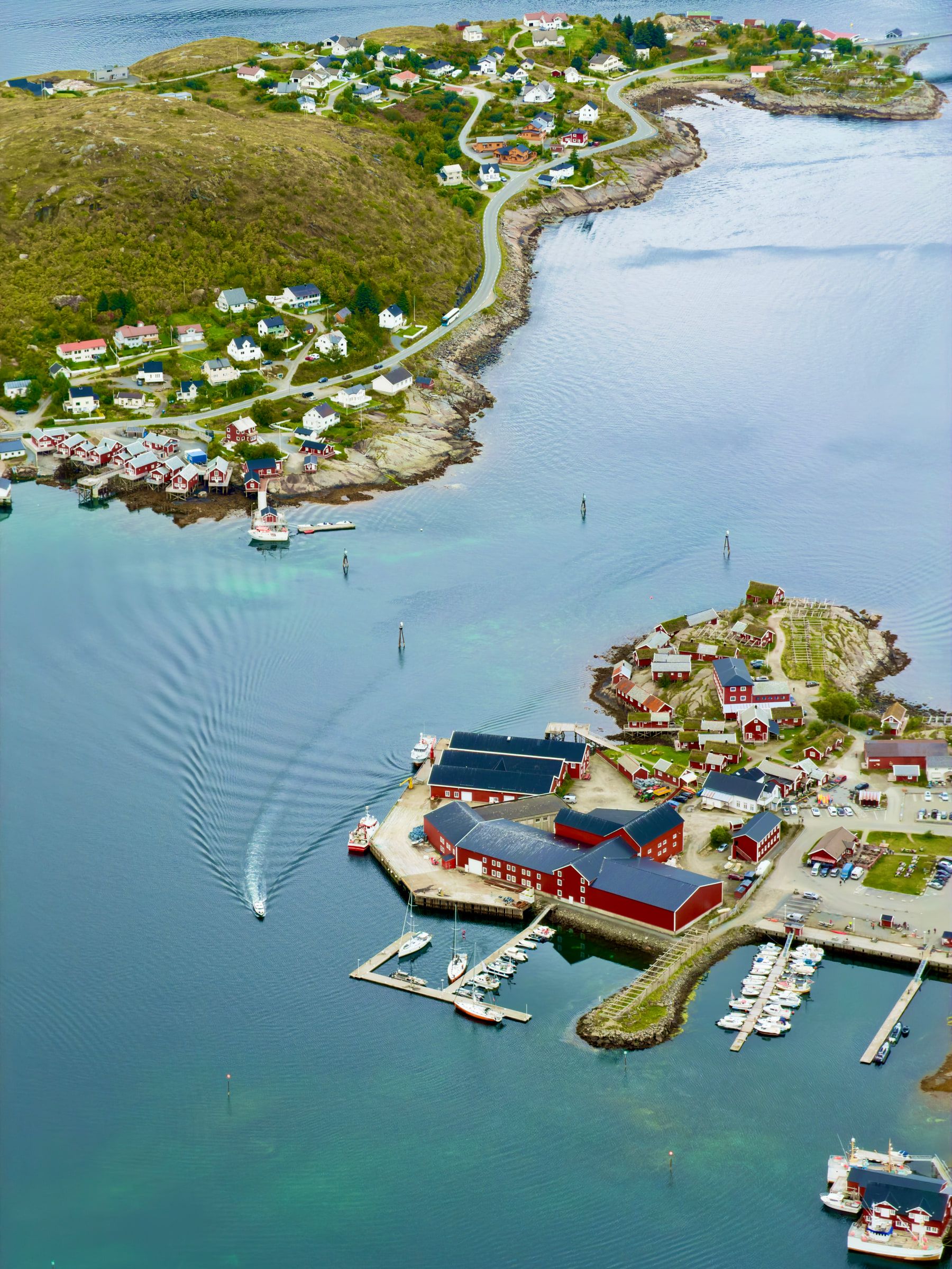 A village of red timber cabins and a small marina on a green headland, seen from high above, with a boat wake crossing the pale water
