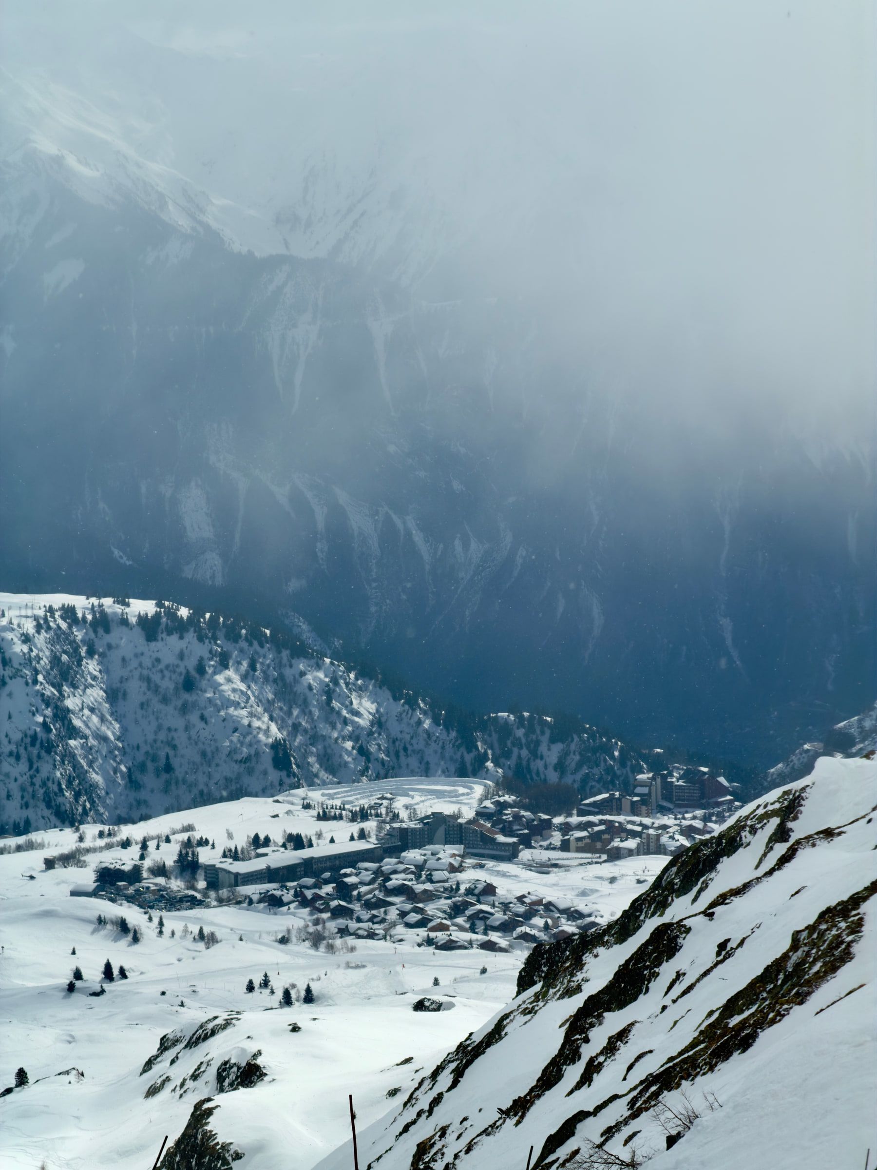 A resort town spread across a snowy bowl far below, with cloud hanging over the mountain wall behind it