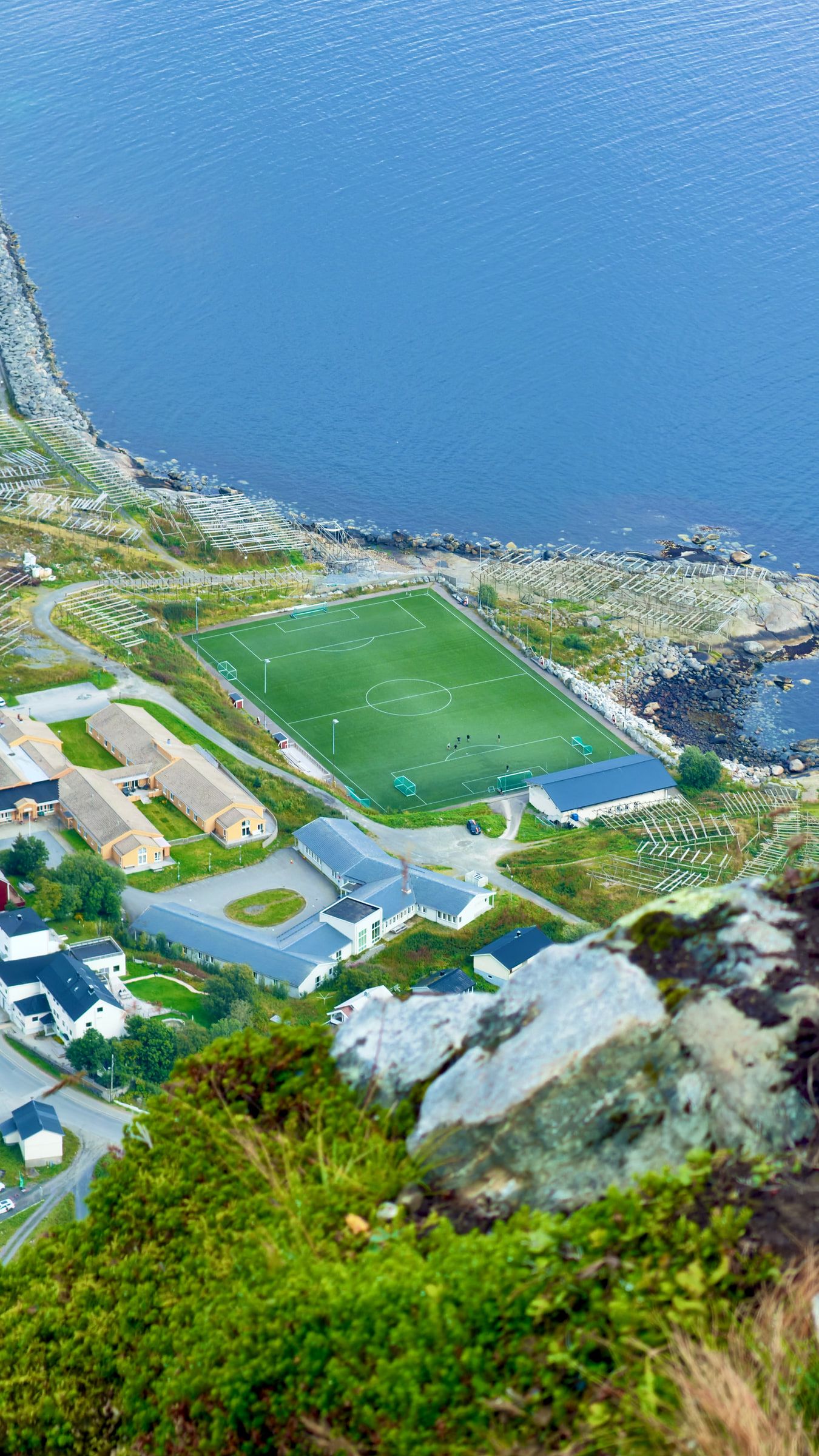 A full-size green football pitch wedged between houses and the shoreline, seen from the ridge above