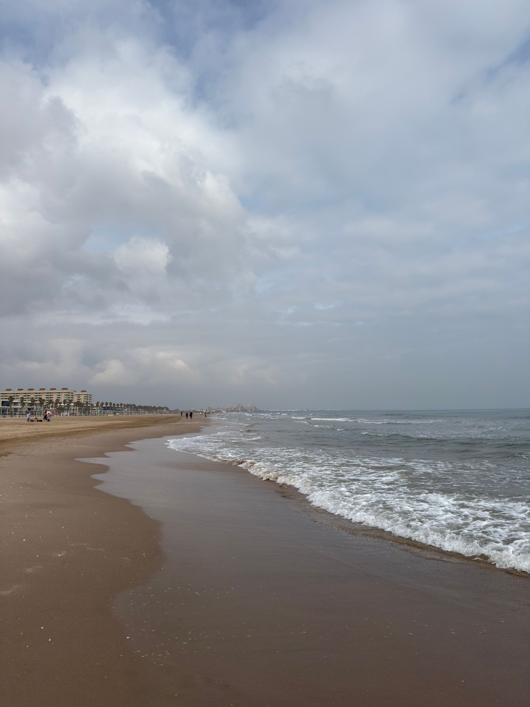 A long flat beach with surf breaking along a curving shoreline under heavy grey and white cloud, a few distant figures walking the sand