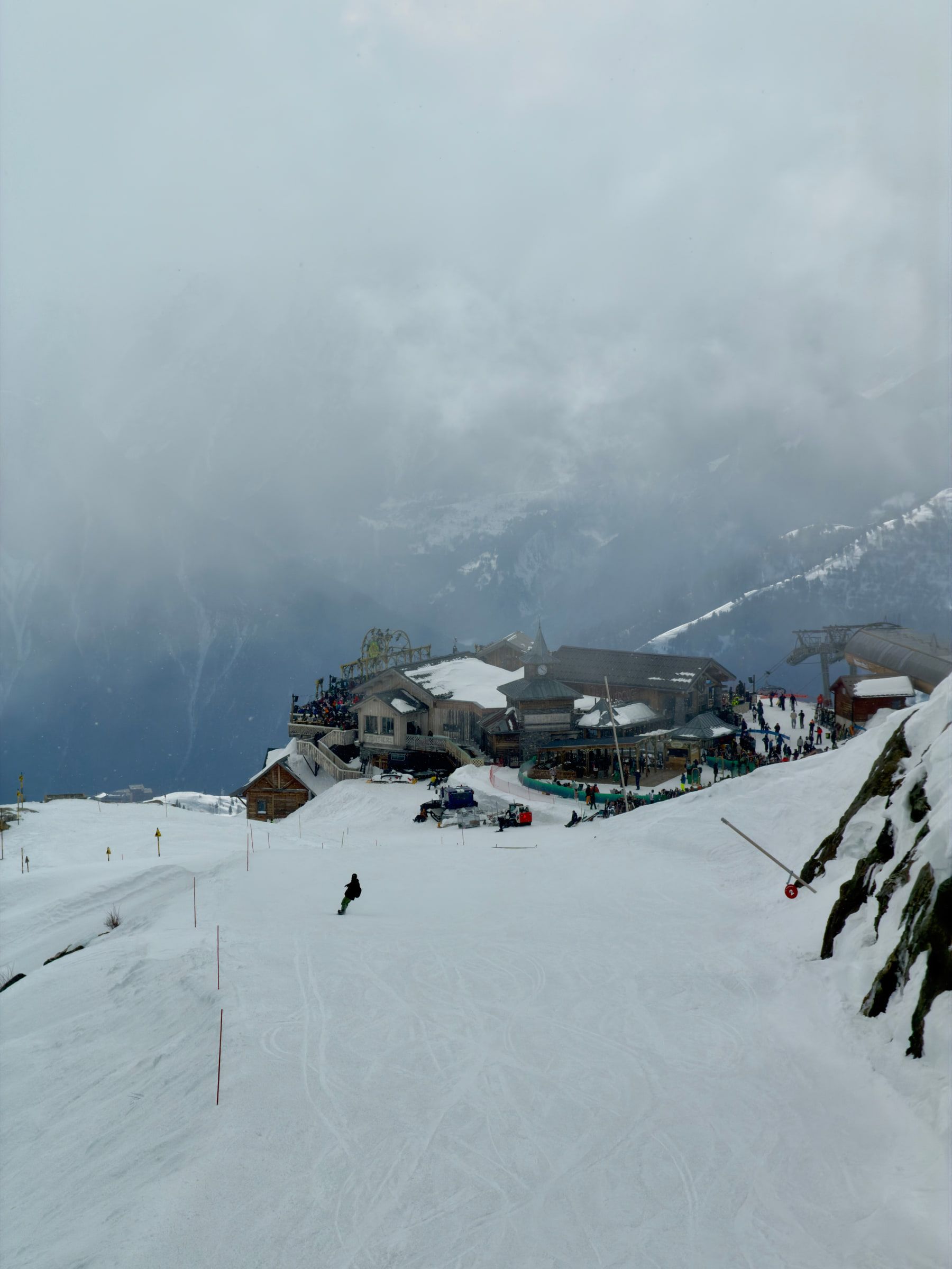 A mountain restaurant terrace built into the piste under low cloud, a single skier passing below it