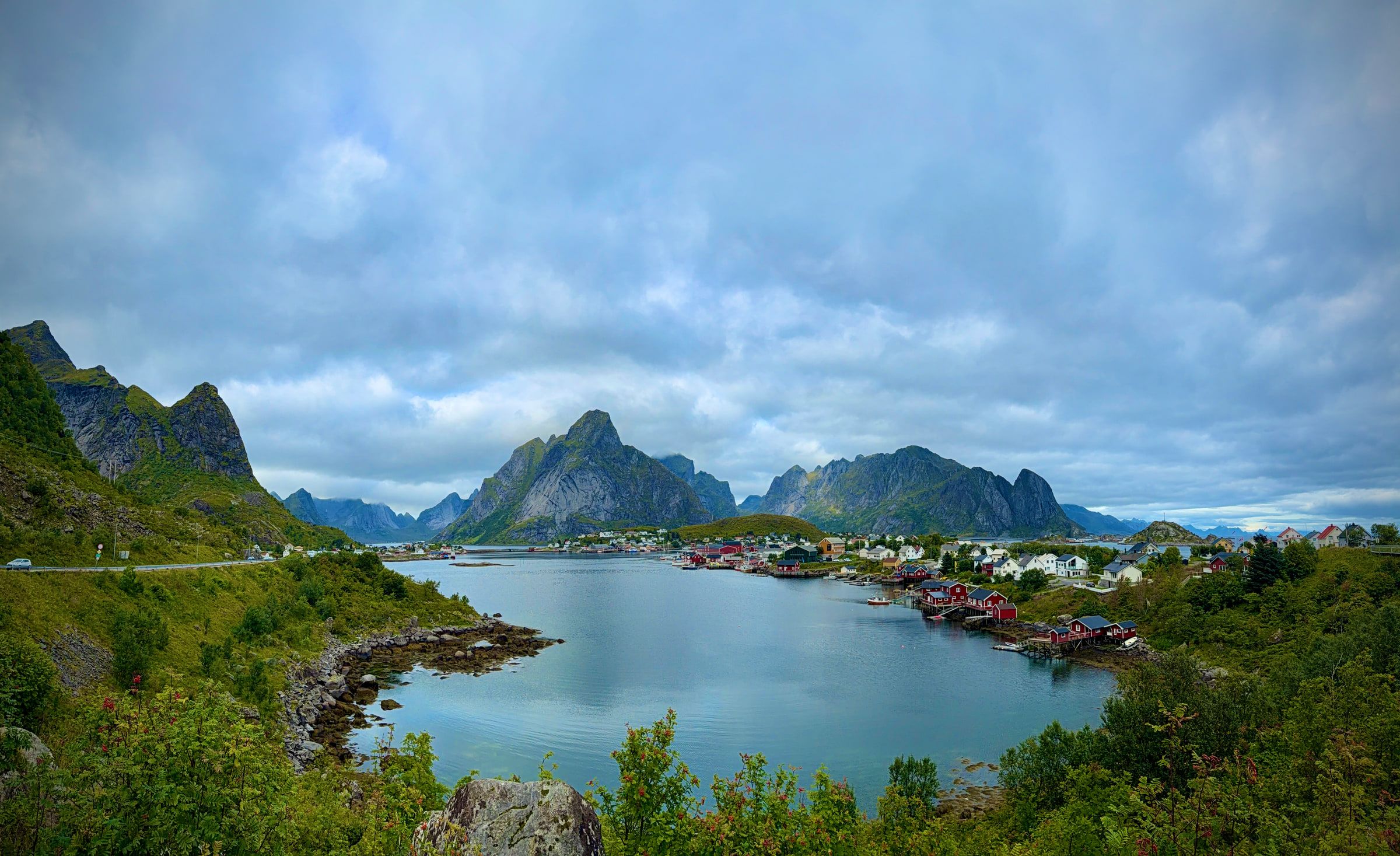 A wide view of a village spread along the water beneath a horizon of jagged grey peaks under heavy cloud