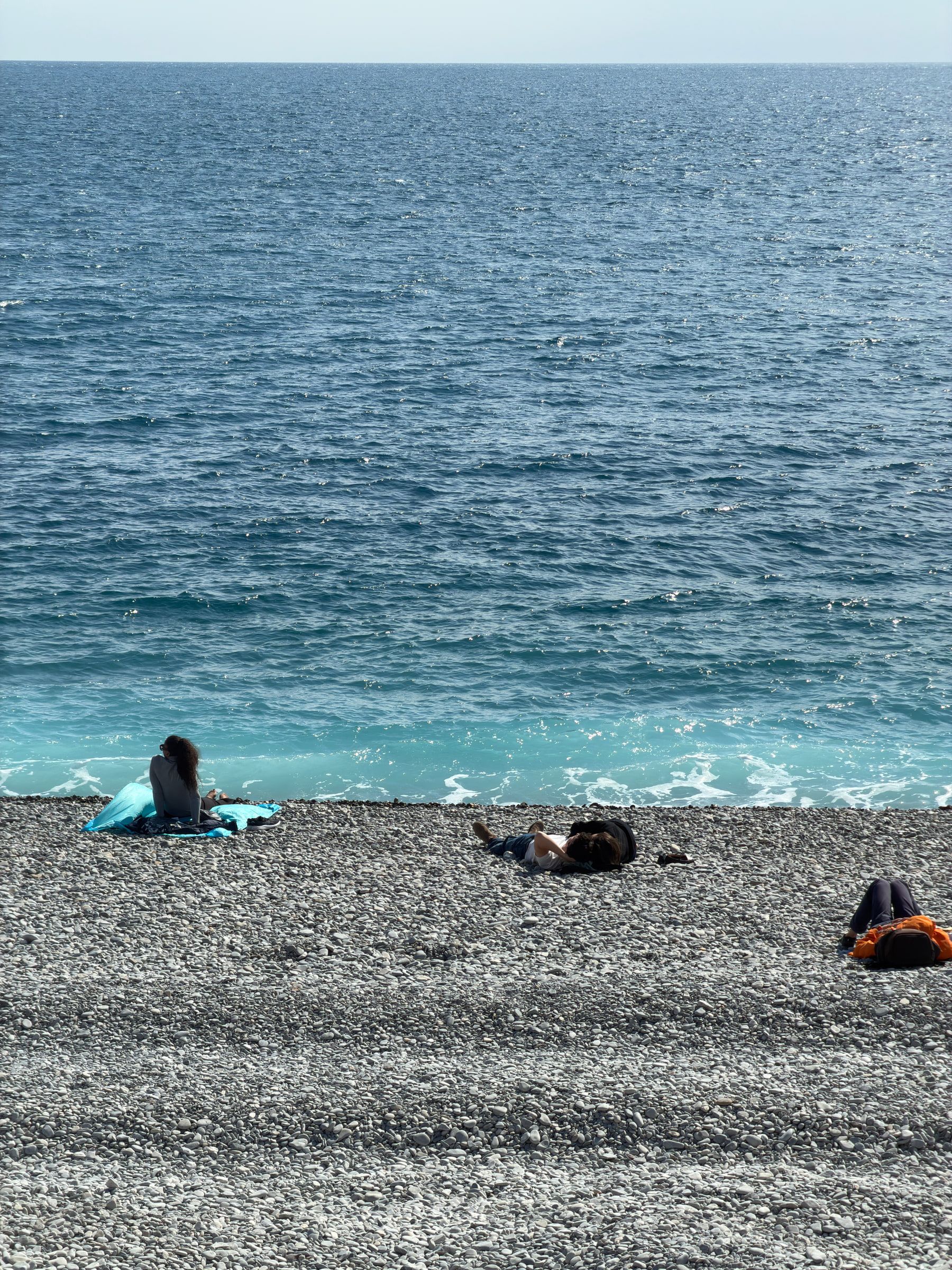 A grey pebble beach meeting turquoise Mediterranean water, a few people lying out on the stones