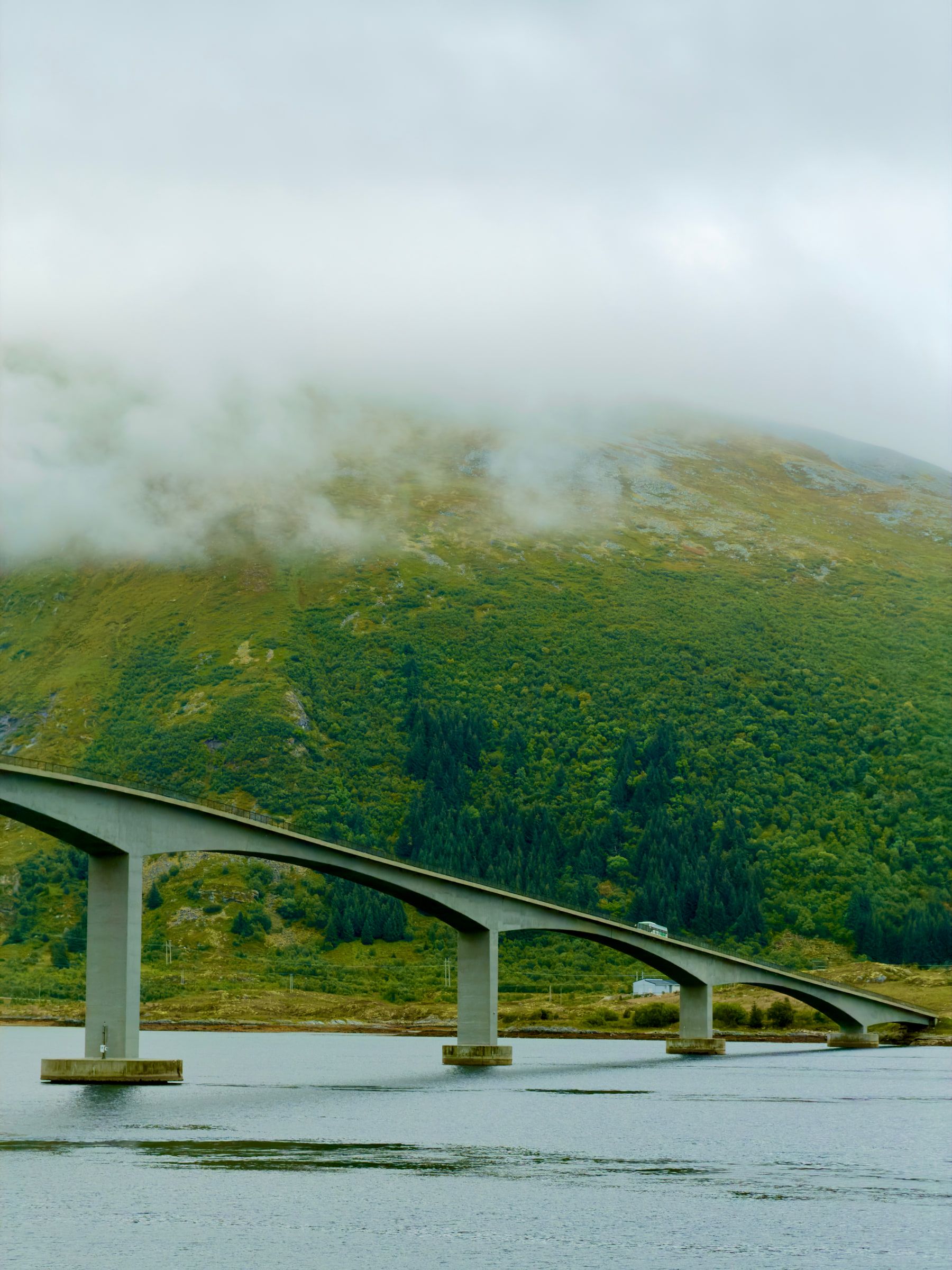 A long low concrete bridge curving toward a green hillside with cloud resting on its summit