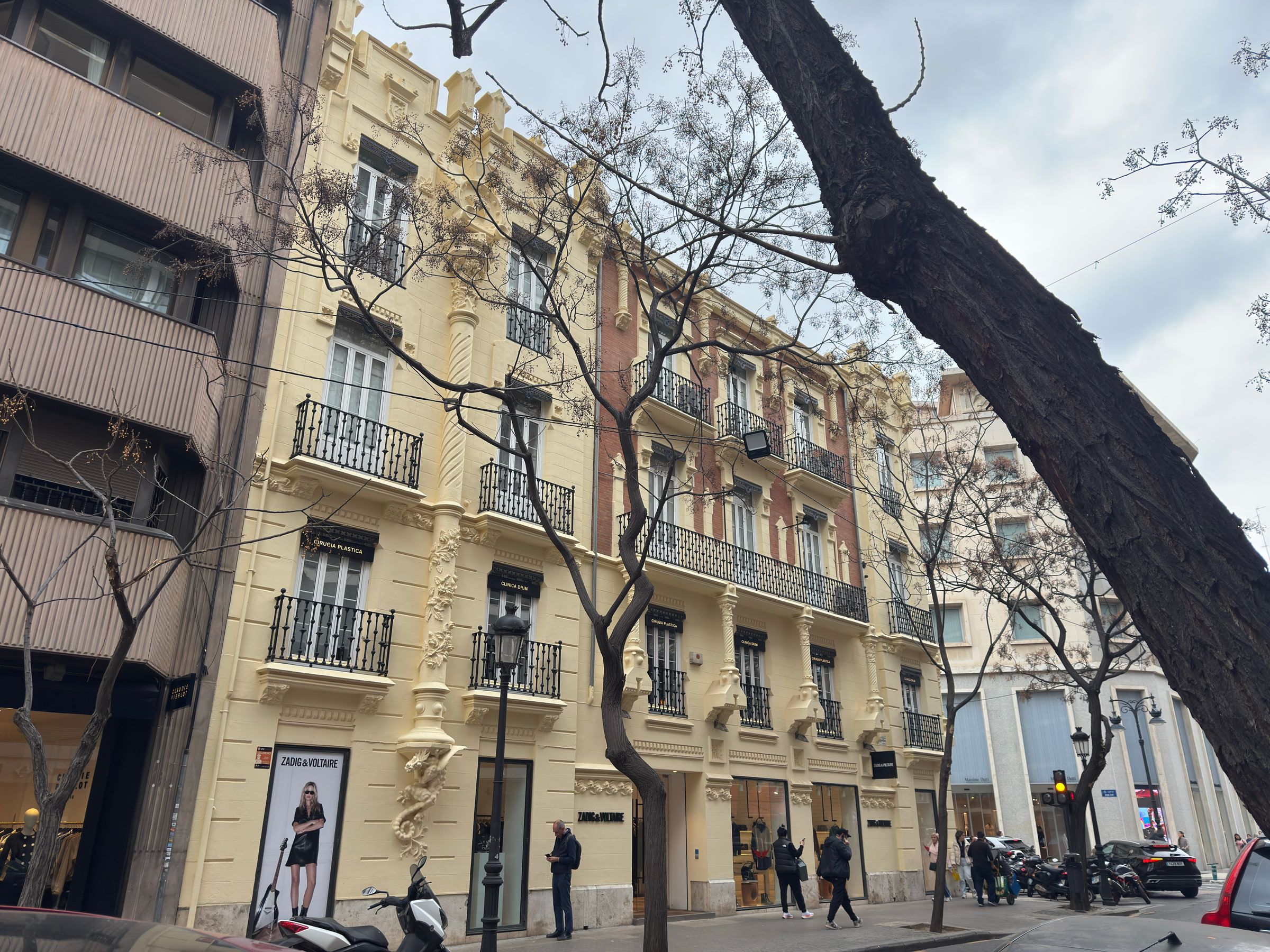 A cream apartment building with carved stonework and rows of iron balconies, bare plane trees in front and people on the pavement below