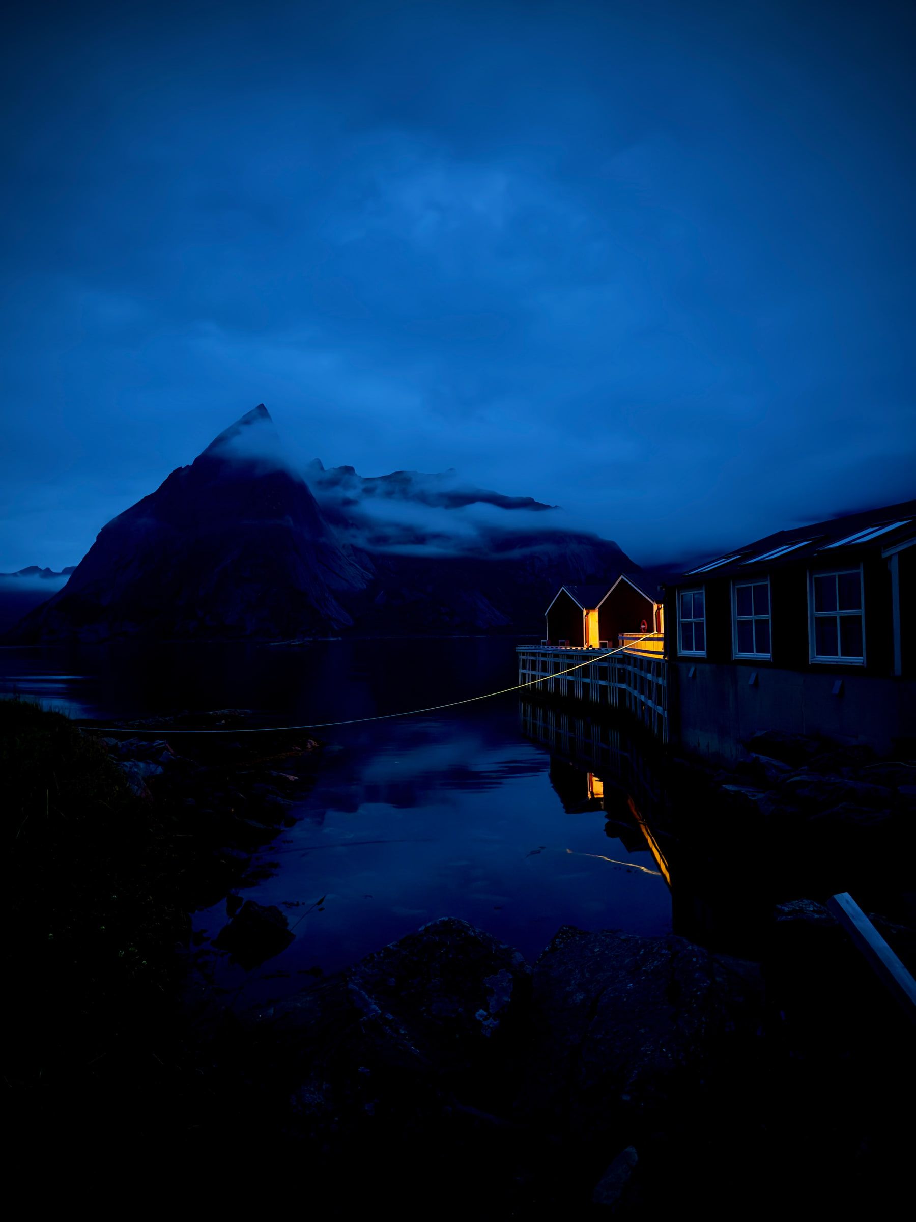 Dark timber cabins on stilts over water at blue hour, their windows lit warm against a silhouetted mountain
