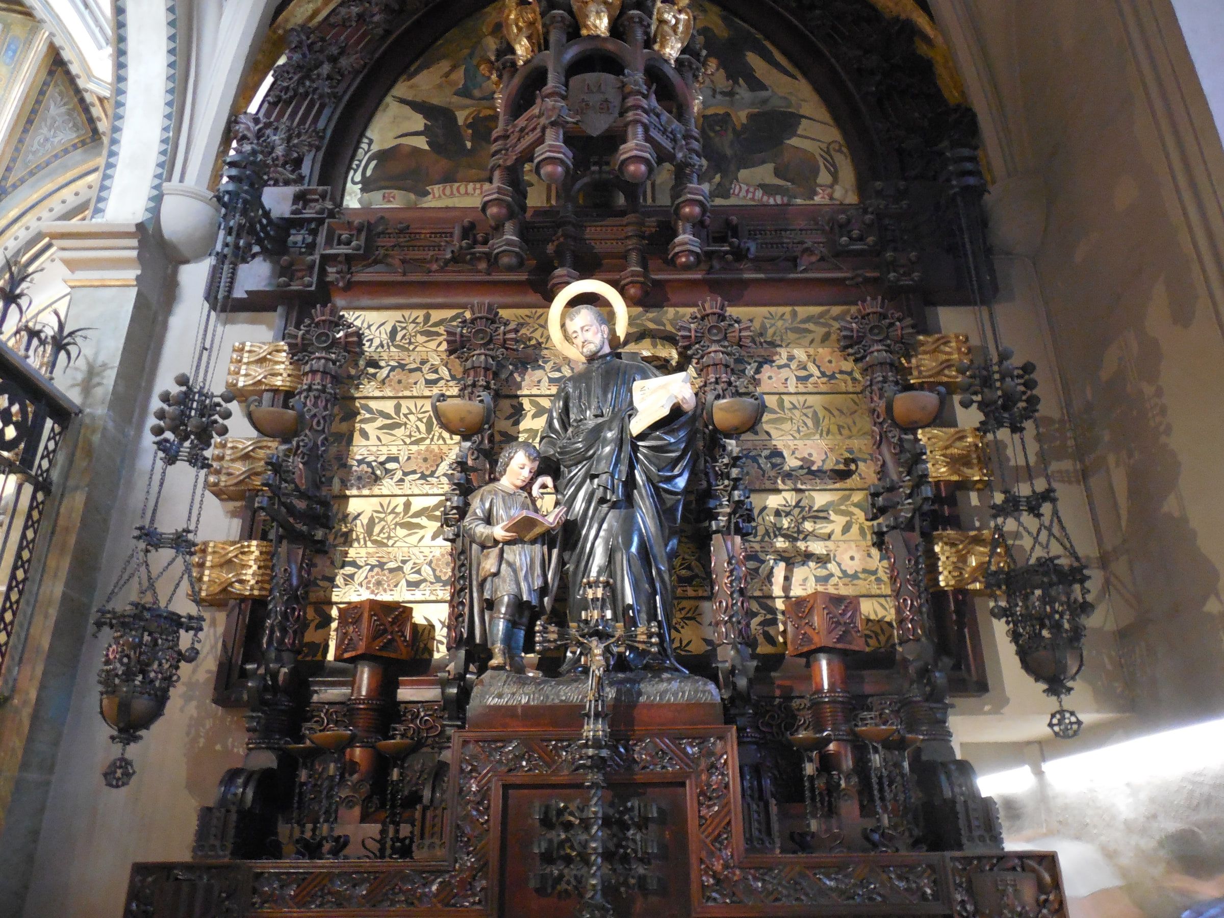 An ornate gilded shrine with a dark-robed standing figure at its centre, wrought ironwork in front