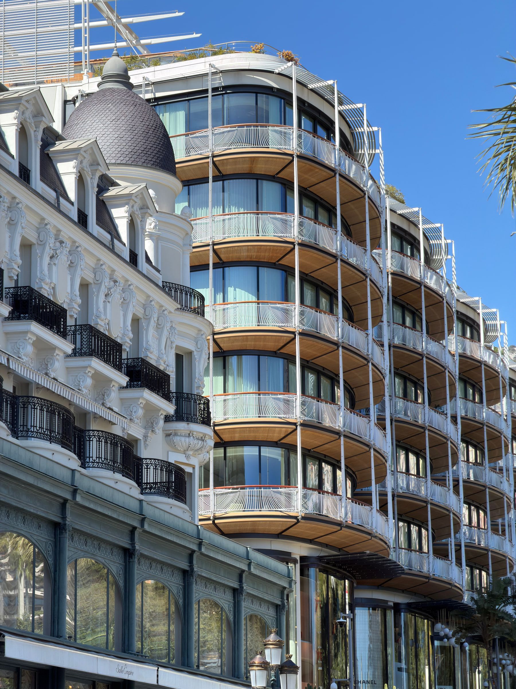 A grand white Belle Époque hotel facade with a dome and tiers of curved balconies, a palm frond crossing the blue sky above