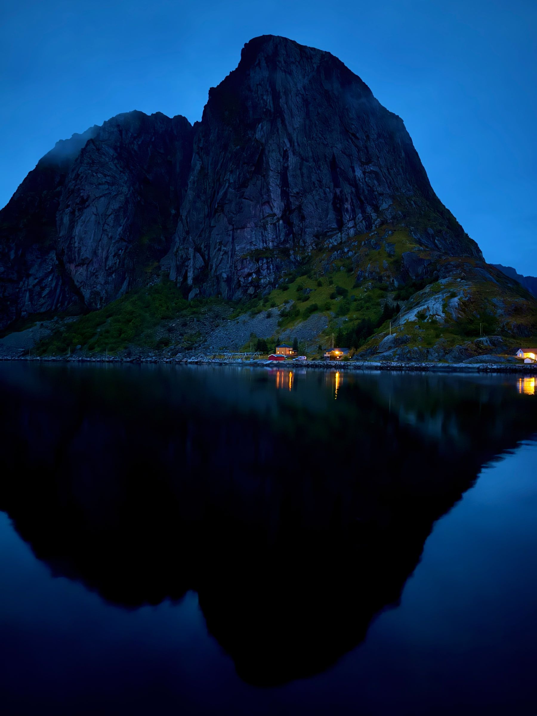 A large dark peak mirrored almost perfectly in still black water at blue hour, small lights along the shore beneath it