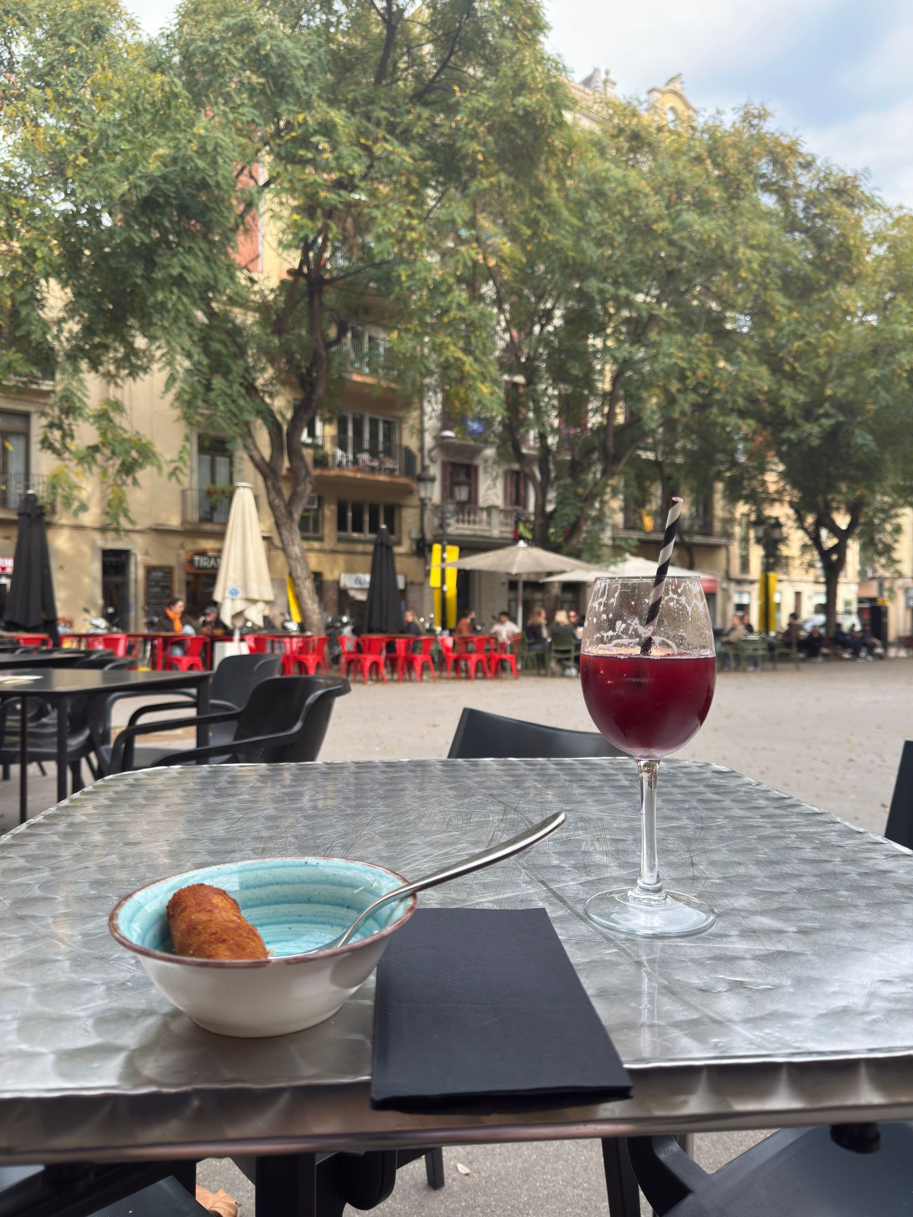 A tall glass of red vermouth with a striped straw and a single croqueta in a small bowl on a metal cafe table, a tree-lined plaza of red chairs behind