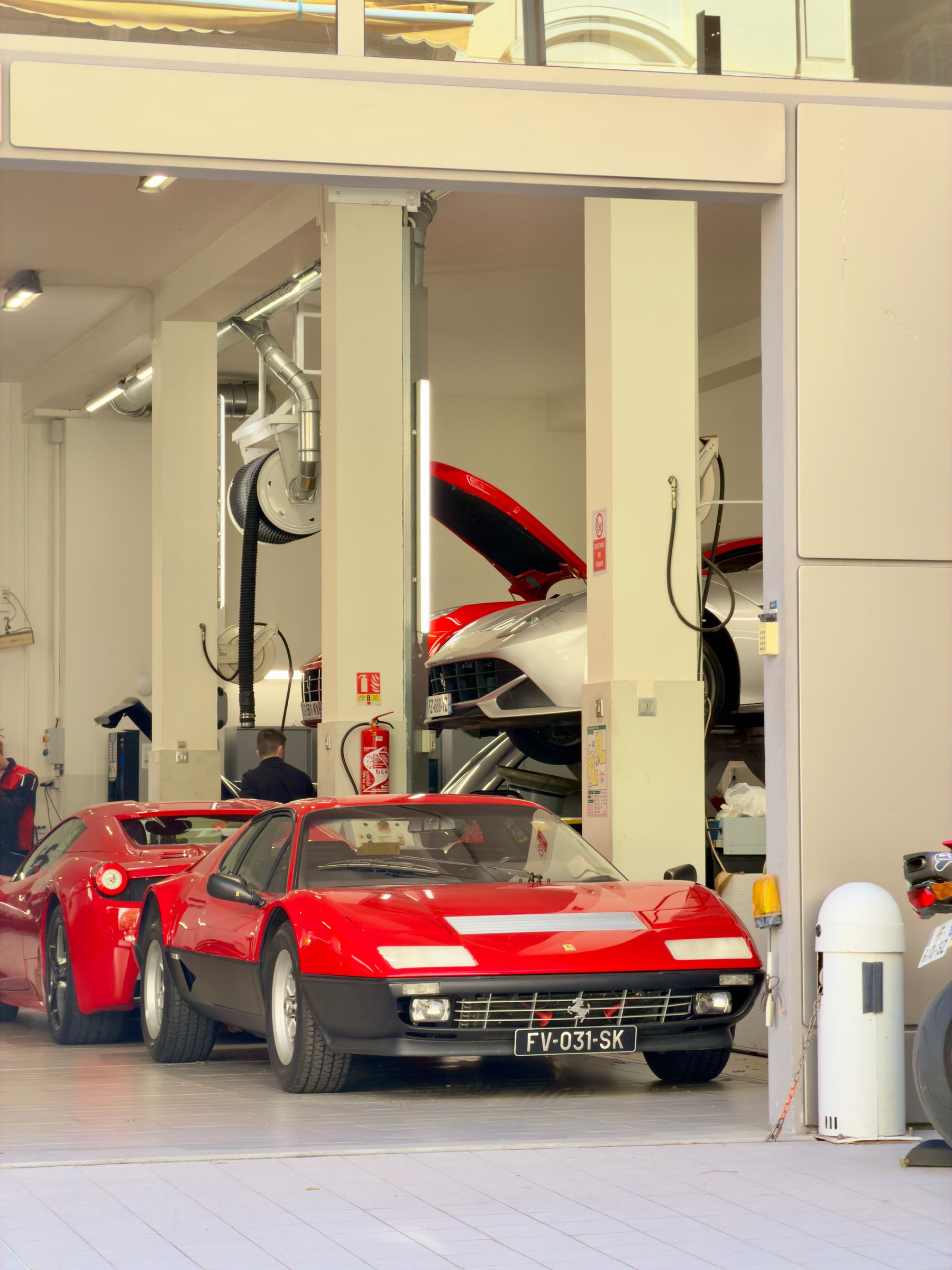 Two classic red sports cars parked in a bright showroom garage, one with its engine cover raised