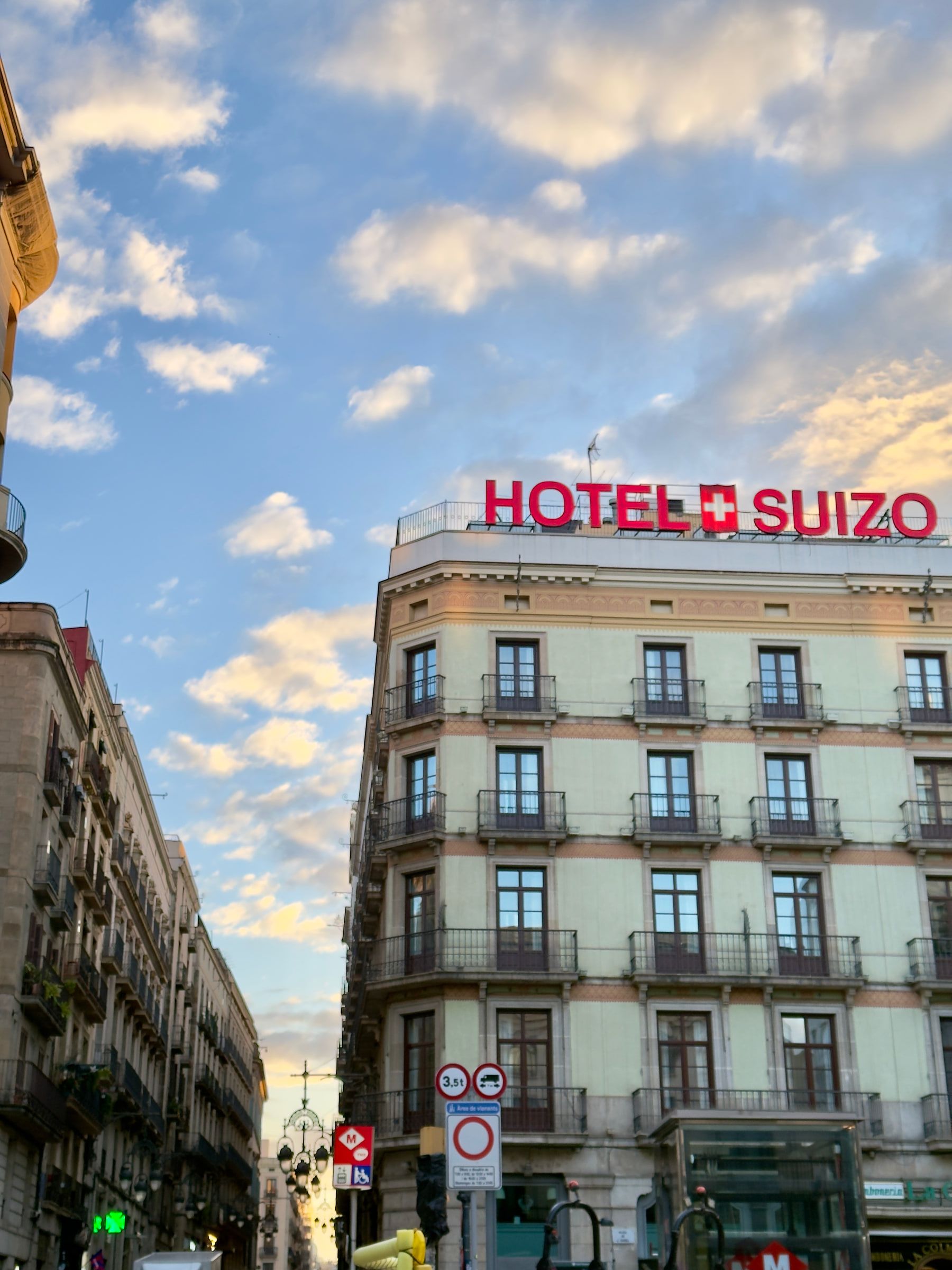 A rooftop sign reading Hotel Suizo above a pale apartment block, lit gold by low sun under a sky of small clouds